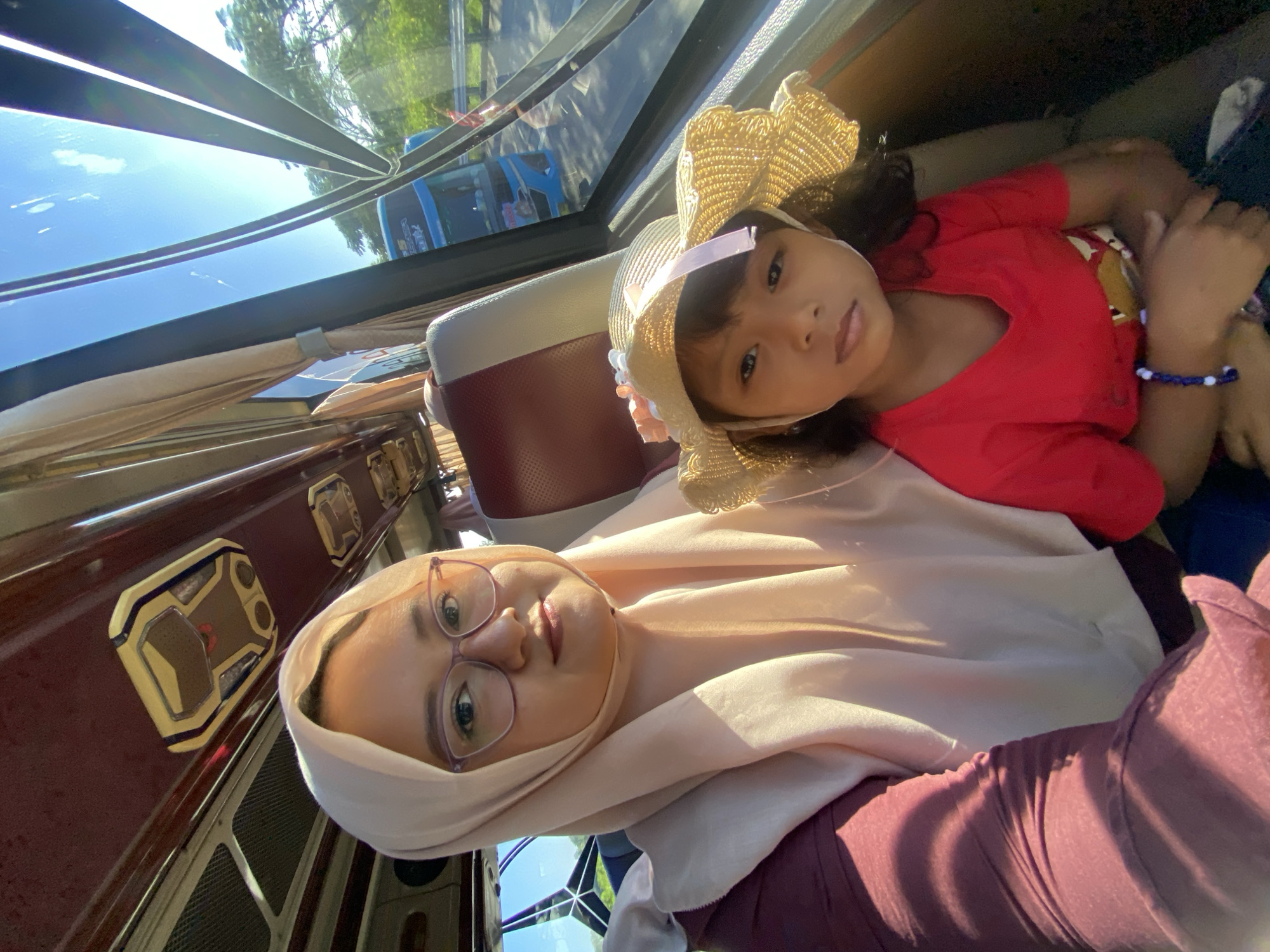 This image captures a selfie inside a passenger vehicle, likely a bus, taken in Mojotengah, Indonesia. The frame is dominated by two individuals: an adult woman on the left and a young girl to her right.
The adult woman, positioned in the foreground, has light skin and wears a light peach-colored hijab that covers her hair and drapes over her chest. She wears light purple-framed eyeglasses and a maroon long-sleeved top. Her gaze is directed towards the camera with a neutral expression.
Seated beside her, slightly in front, is a young girl with a darker skin tone. She wears a red short-sleeved top and a straw hat adorned with a light pink ribbon and small white floral embellishments on the left side. A blue and white beaded bracelet is visible on her left wrist. She also looks directly at the camera with a neutral expression.
The interior of the vehicle features red and white two-tone seats. Overhead, a maroon ceiling panel is visible, equipped with rectangular light fixtures or vents, one featuring a golden and dark brown design. A large window on the right side of the frame reveals the exterior environment. Light-colored curtains are pulled back on the left side of the window.
Through the window, a paved road is visible, alongside a metal guardrail. A blue and white passenger bus, partially visible, is on the road, with "TRANS BUS" or similar text discernible on its side. Beyond the road, green trees and foliage are present under a clear blue sky. Strong natural sunlight enters from the right side of the vehicle, casting bright highlights on the subjects and interior.