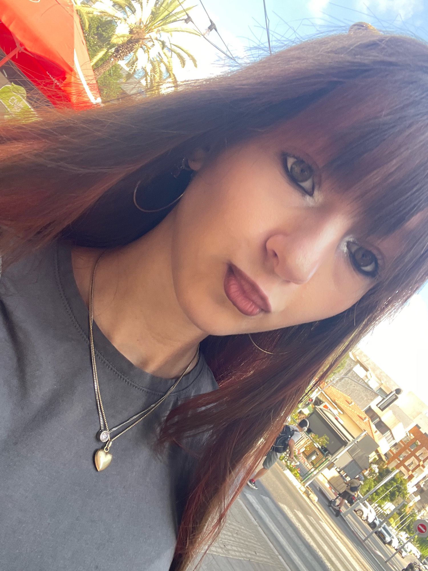 A young woman with long, auburn hair and bangs takes a selfie outdoors, with a cityscape visible in the background. She is wearing a dark grey t-shirt and a gold necklace with a heart-shaped pendant. The environment appears to be a street in Raanana, Israel, with palm trees, buildings, and traffic visible. The woman's facial expression is neutral.