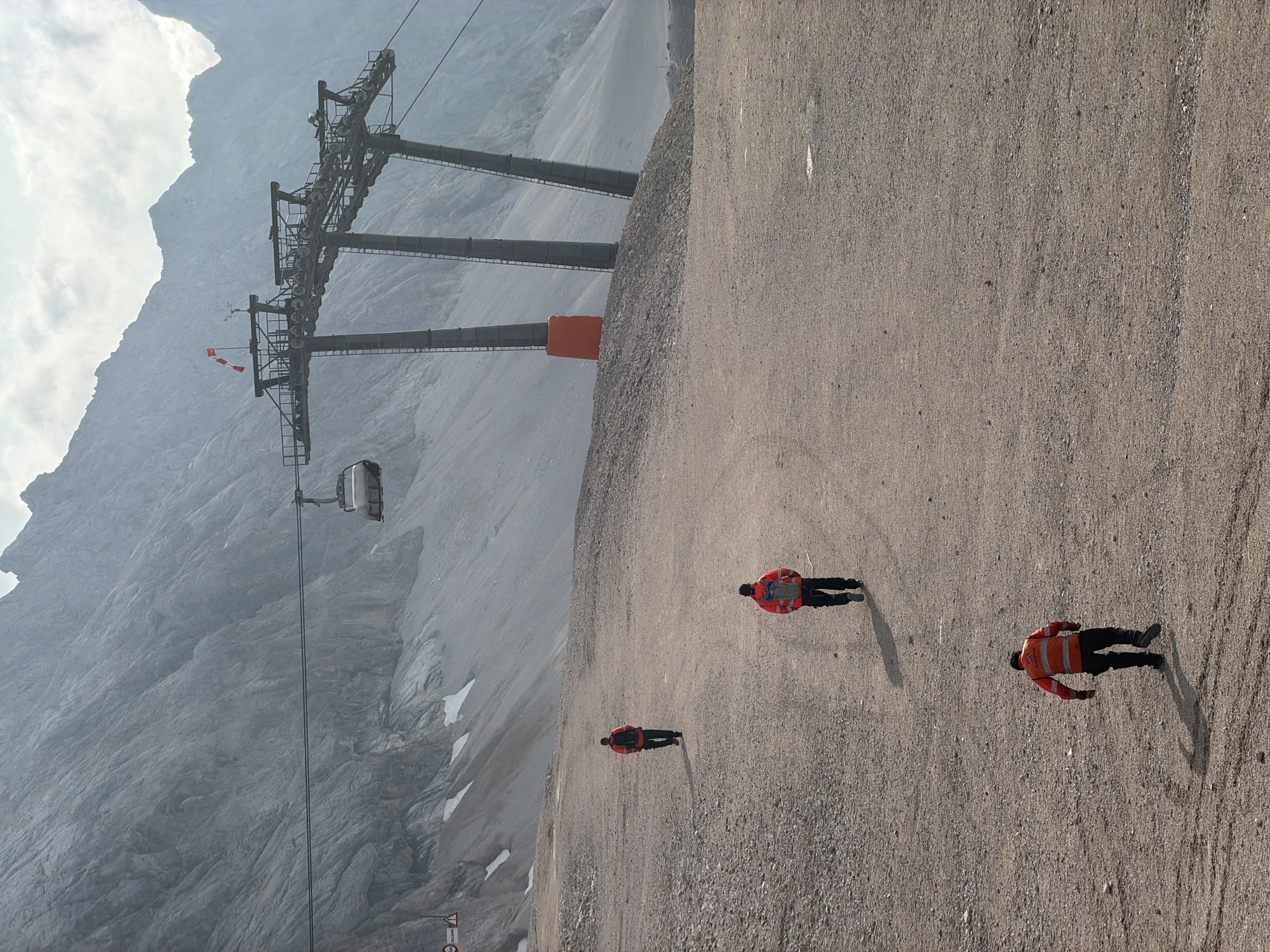 The image depicts a mountain landscape with a cable car system in the background, likely at the Zugspitze, Germany's highest peak, near Garmisch-Partenkirchen. Three individuals, dressed in reflective orange vests and dark pants, are walking away from the camera on a gravelly path. One individual is closer to the foreground, with two others positioned further up the path and slightly to the left. The cable car station with its support towers and a suspended gondola is visible on the slope of the mountain. The terrain is rocky and barren, with patches of snow or ice scattered on the mountainside. The sky is overcast with some clouds.
