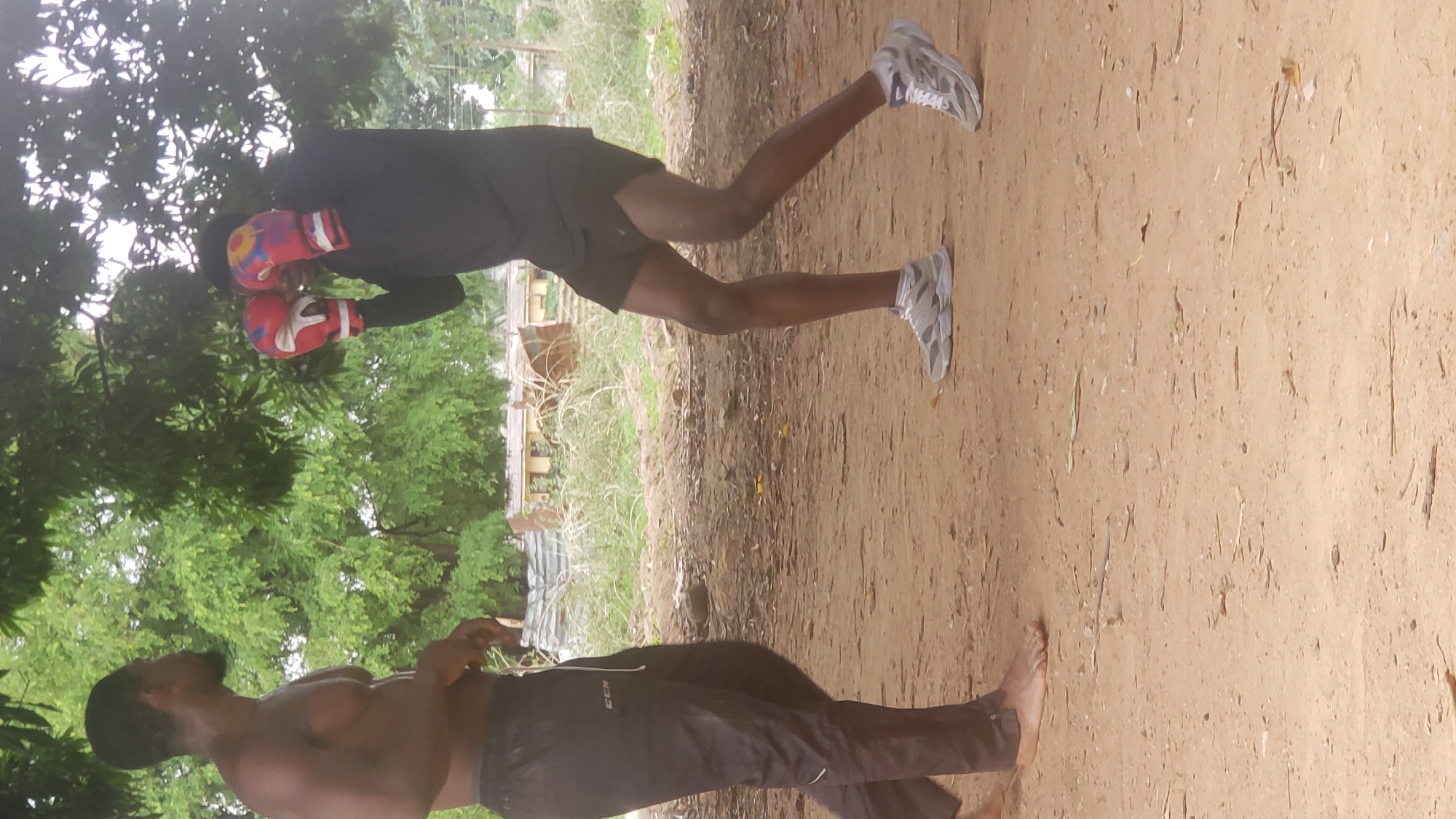 The scene is outdoors, likely in a training area in Narayi, Nigeria. A shirtless man with dark hair and a beard stands to the left. He is wearing dark-colored pants. Another man is wearing a long-sleeved black shirt, dark shorts, and grey sneakers. He is wearing red and yellow boxing gloves and appears to be engaged in boxing training. The ground is dirt and dry grass. Green trees border the area.