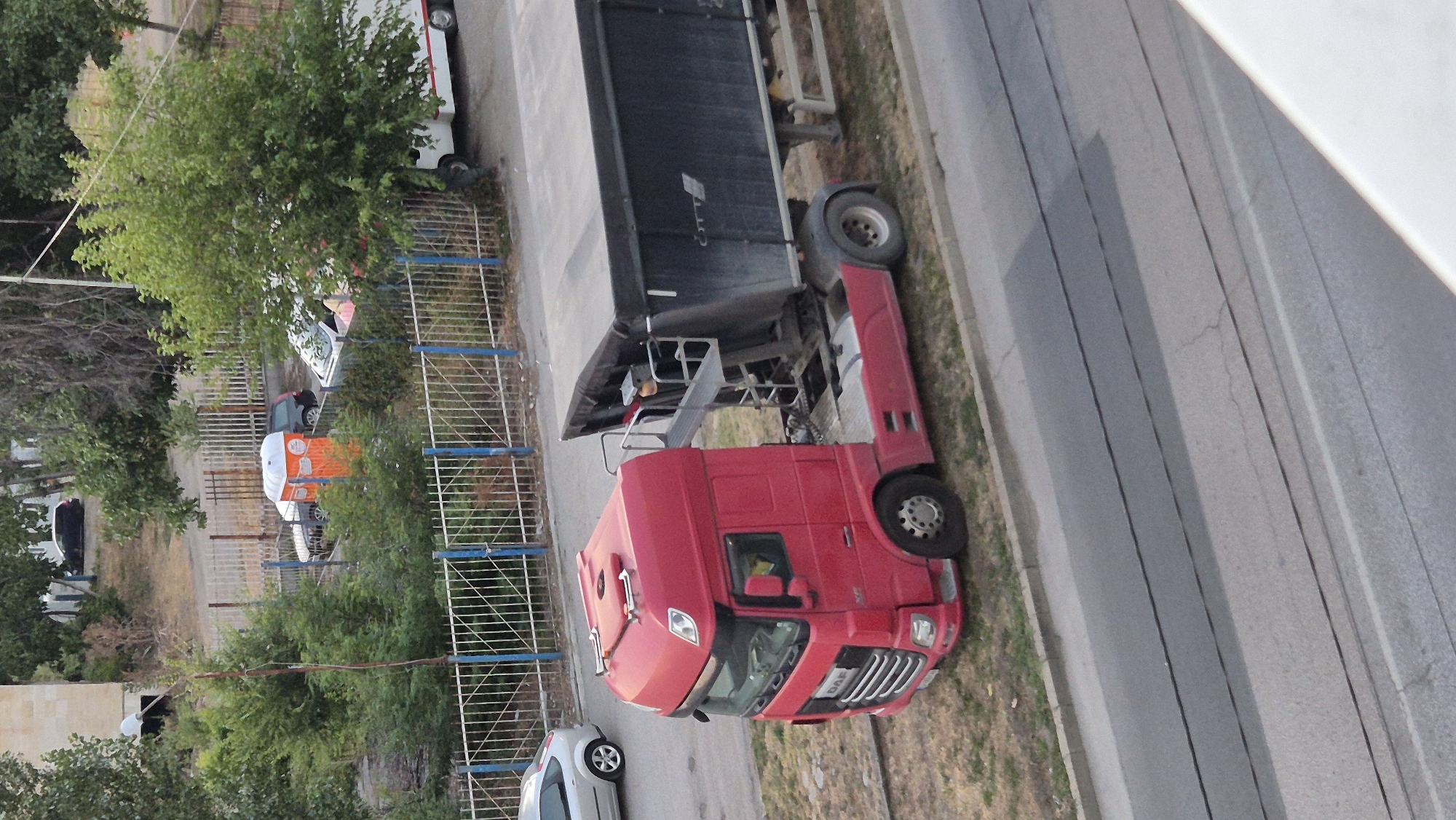 A red DAF truck with a black tarp-covered trailer is parked on a grassy shoulder next to a road. The truck is facing left, and the trailer is behind it. The trailer appears to be a dump trailer. In the background, there is a white van, a portable toilet, and several cars parked behind a fence. There are trees and buildings in the background. The scene is likely in Lovech, Bulgaria, given the context.