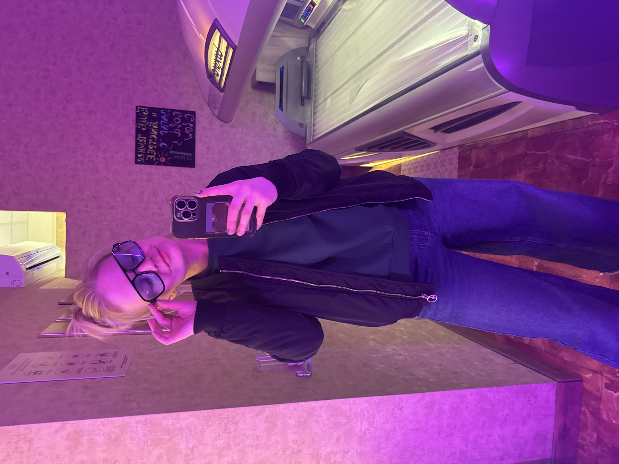 A person takes a selfie in a tanning salon. The individual, likely a woman with fair hair, wears sunglasses, a black jacket, a dark green top, and blue jeans. She holds a smartphone with a multi-lens camera. The tanning bed is visible in the background, along with a sign in Russian. The purple lighting suggests an artificial setting typical of tanning facilities. The location is likely in Saint Petersburg, Russia, considering the use of the Russian language.