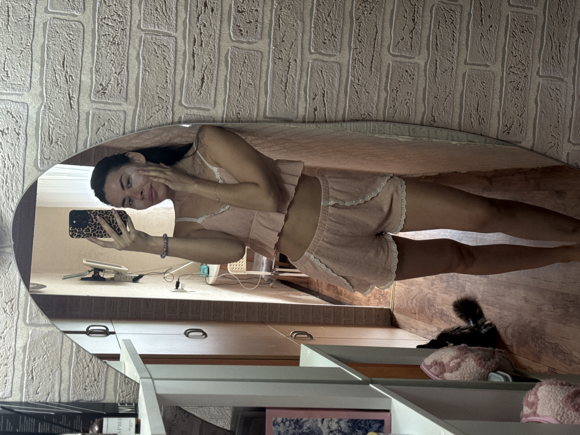 The media file shows a young woman taking a mirror selfie. She is wearing a pink pajama set with lace trim and has a facial mask applied to her cheeks. The woman is holding a smartphone with a leopard-print case in her left hand, and her right hand is touching her face. She has dark hair and is looking at her reflection in the mirror.
The setting appears to be a bedroom or dressing area, with a textured brick-patterned wallpaper visible on the wall in the reflection. A wooden floor is also visible, along with some furniture, including cabinets with handles. A cat is partially visible lying on a shelf in the lower right corner of the image.
The image is a vertical shot, and the lighting suggests it might be daytime, though no specific time can be determined. The overall mood seems relaxed and casual, consistent with being at home in pajamas. There is no visible text in the image. The location context is Chaikovsky, Russia.