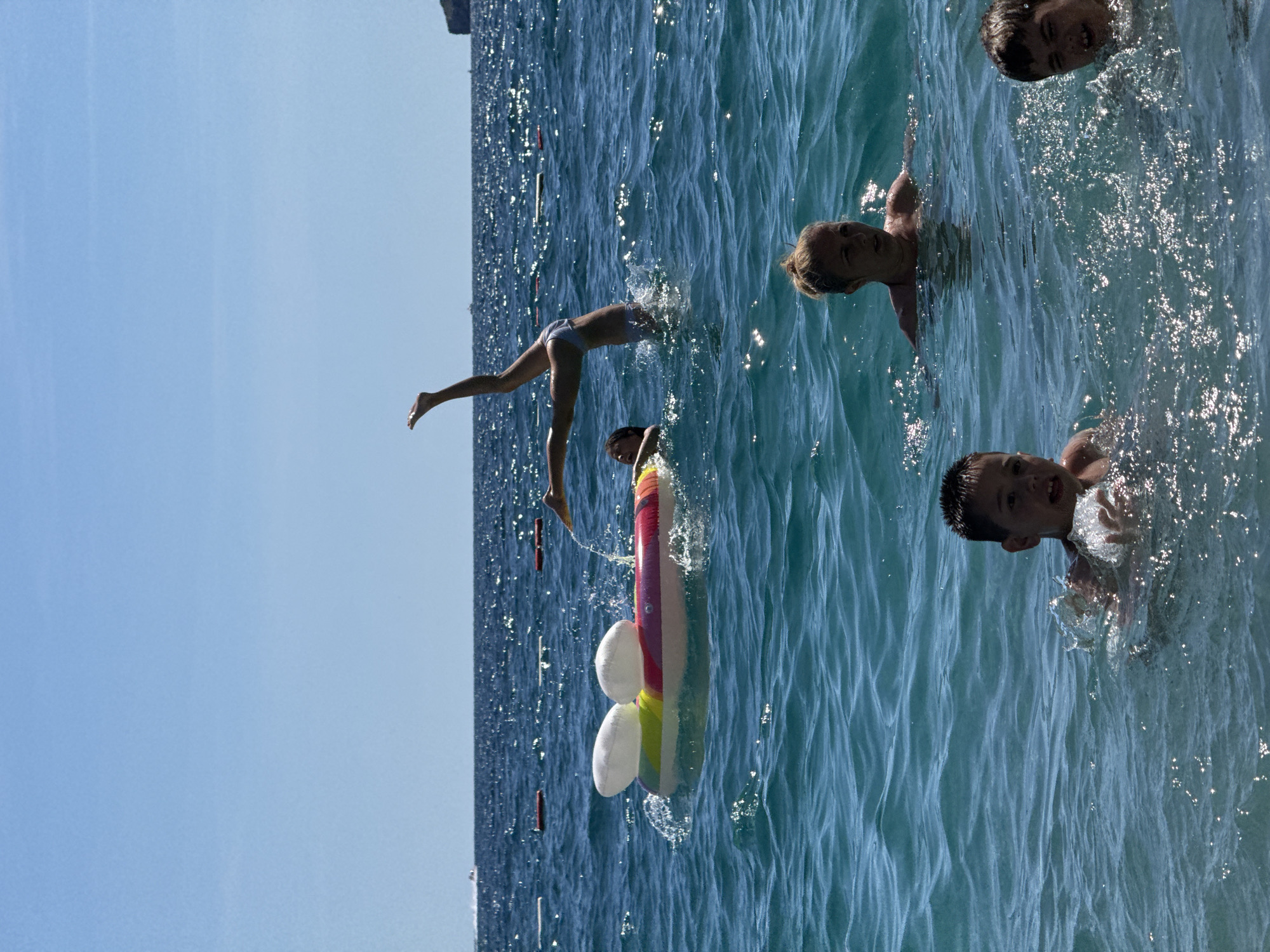 In the city of Kamenovo, Montenegro, on a bright, sunny day, several individuals are enjoying the clear blue water of the Adriatic Sea. In the foreground, two young boys are swimming, with one splashing water. Behind them, a woman with blonde hair tied up in a bun is looking forward, appearing to be in the water. Further back, a person is performing a handstand with their legs in the air. Adjacent to this person is an inflatable raft shaped like Mickey Mouse's head, with white ears and a colorful body. The sea is calm with gentle waves, and the sun reflects off the water's surface. In the distance, a small boat is visible on the horizon, and a rocky coastline can be seen to the right. Red and white buoys mark a boundary in the water.