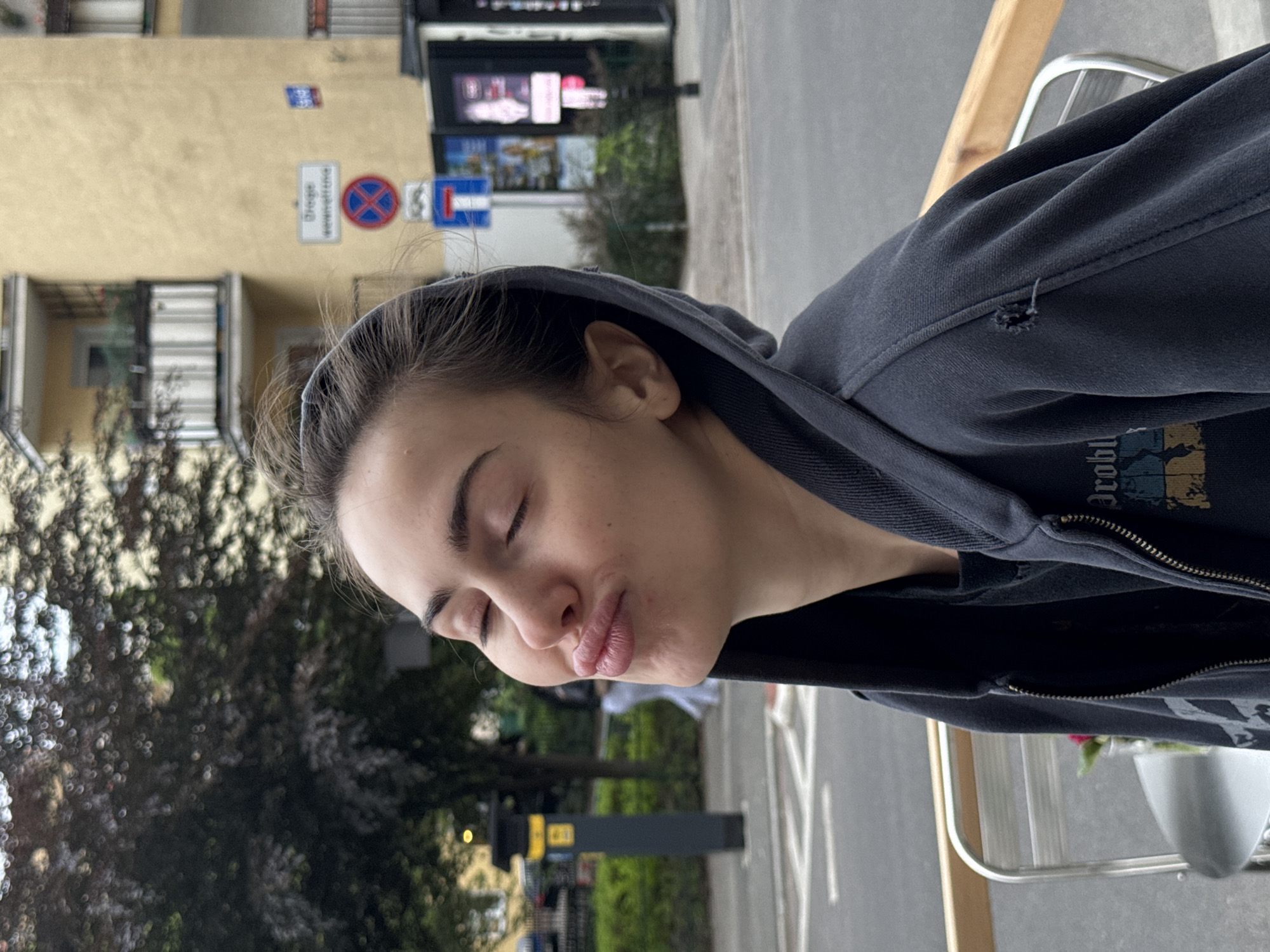 A close-up, portrait-oriented shot captures a young person from the chest up, positioned slightly off-center to the left. The person has fair skin, dark hair pulled back, and their eyes are closed while their lips are pursed in a playful "kissing" or "pouty" expression. They are wearing a dark grey or black hooded sweatshirt with a zipper partially visible. A small, ragged tear or hole is visible on the sleeve of the hoodie near the shoulder. Partial text, possibly "Dro...bl", can be seen on the front of the sweatshirt.
The background suggests an urban street scene under what appears to be an overcast sky during the daytime. To the left of the person's head, a light-colored building with multiple windows and balconies is visible. In front of the building, several street signs are mounted on a pole. One white rectangular sign prominently displays German text, partially visible as "Ortsge... wenn ni...t", which translates to "Urban area unless otherwise ordered". Below it is a red circular sign with a blue X, indicating a "no stopping" or "no waiting" zone, next to a blue rectangular sign with a white arrow pointing right, likely signifying a mandatory direction or one-way street. Further down the street, blurred trees, other buildings, and large, illuminated advertising screens are visible. To the right of the person, part of a metallic outdoor chair or railing is present, hinting at an outdoor seating area.