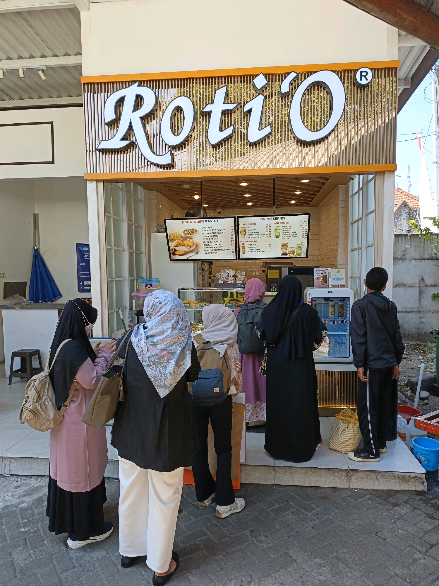 The image shows a street-level view of a Roti'O bakery in Surakarta, Indonesia. A sign above the entrance reads "Roti'O". Several people, predominantly women wearing hijabs, are queuing outside the shop. One person, a young male, stands to the right of the queue. In front of the shop, a refrigerated display case contains drinks. A menu board above the counter lists "SIGNATURES' PASTRY" and "DRINK SERIES" with various item names and prices. The bakery appears to be open for business. The surrounding environment shows a paved walkway, a building facade with windows, and some outdoor elements such as an umbrella and buckets. The sky is visible in the upper right corner.