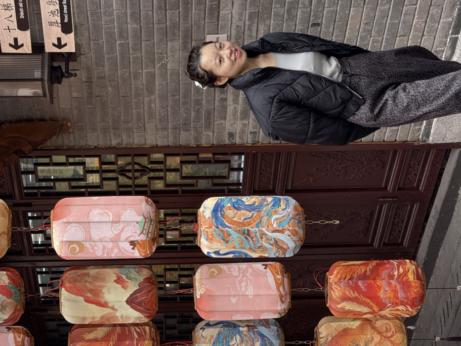 An Asian woman stands near a brick building with hanging signs. Below her is a brown wooden door adorned with carved designs. Rows of rectangular lanterns in various colors and patterns hang in front of the door. The woman wears a black puffer jacket over a light-colored shirt and patterned pants. She is smiling and looking at the camera. The scene is colorful and appears to be taken in an urban setting.