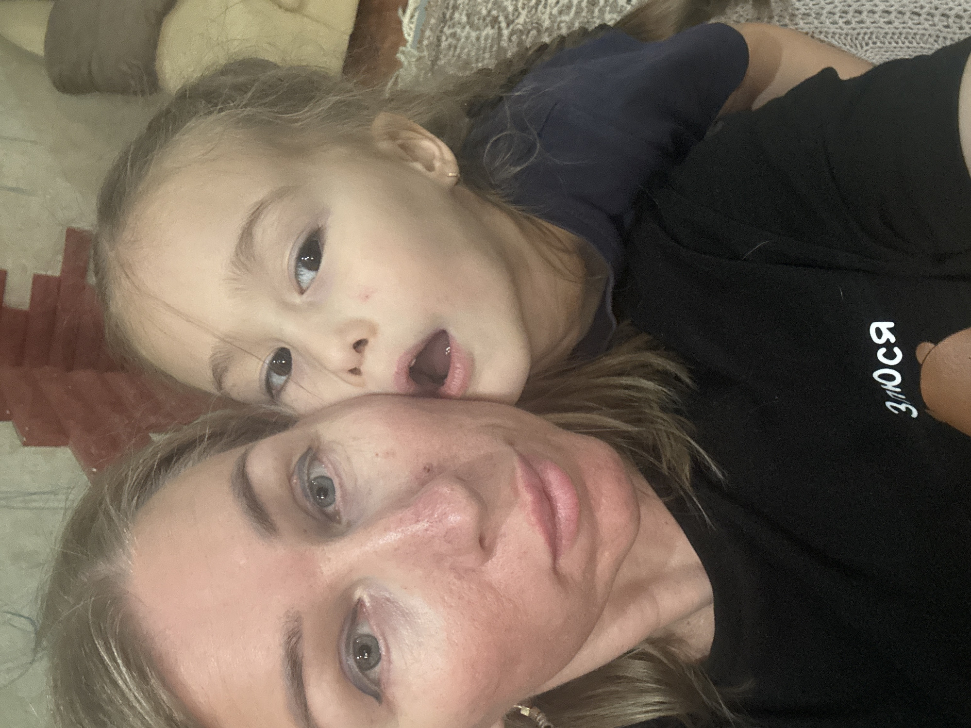 A woman and a child, likely a mother and daughter, are in a close-up selfie. The child has light brown hair and is wearing a dark blue shirt; she has an open mouth expression. The woman has light brown hair and is wearing a black shirt with white Cyrillic letters on the sleeve. The background is somewhat blurred, showing parts of furniture and what appears to be a textured blanket or surface. The location and city of Tashkent, Uzbekistan are not confirmed or suggested by the details in the image.