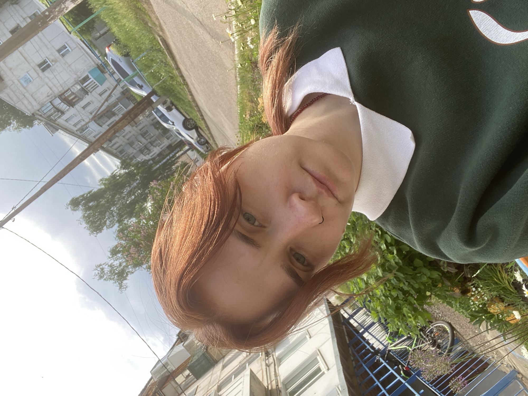 A reddish-haired person wearing a green and white top is in an outdoor setting. The background contains apartment buildings, trees, parked cars, and a sidewalk. A bicycle is also visible near a blue fence. The location is Chaikovsky, Russia.