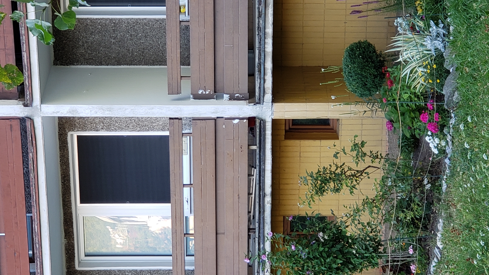 The image shows a close-up of a residential building's exterior, featuring balconies and a ground-level garden. The building appears to be made of concrete and yellow brickwork. The balconies have brown wooden railings. A window with a dark screen is visible on the upper level. The ground-level garden contains various plants and flowers, including pink, white, and magenta blossoms, as well as green shrubs and a neatly trimmed spherical bush. A wire fence runs through the garden. The scene is set outdoors, likely in a city environment, as suggested by the building's architecture. The photograph is taken from a slightly low angle, looking upwards at the building.