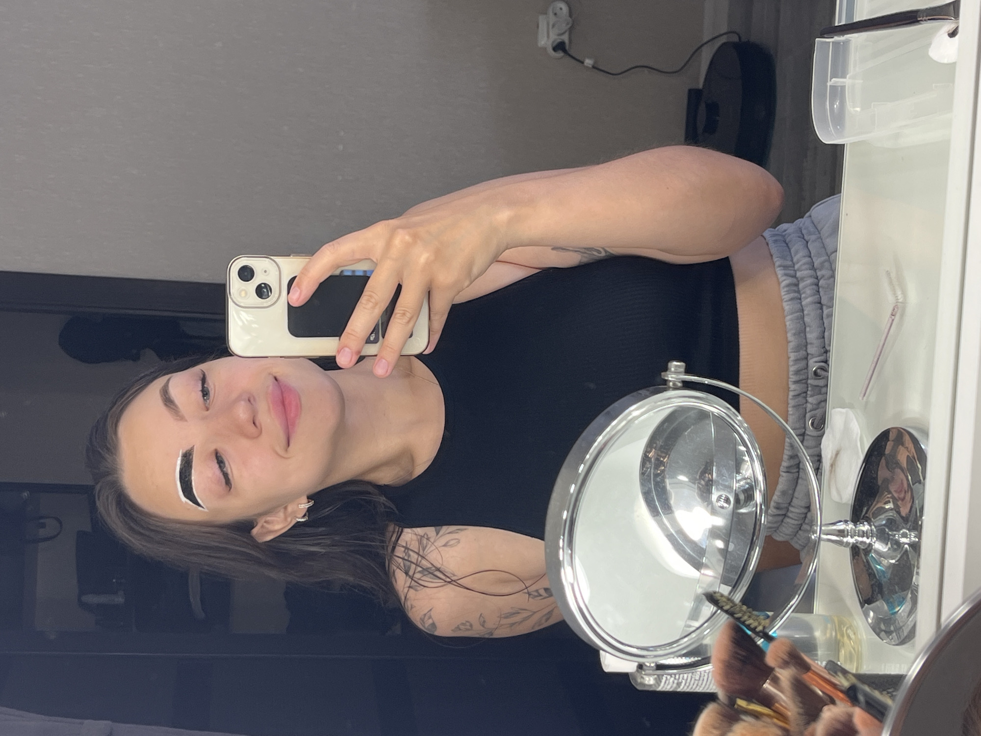 A young woman is taking a selfie in a mirror, with a phone held in her right hand. She is wearing a black tank top and grey sweatpants. Her left eyebrow has a white substance applied to it. There are makeup brushes and a mirror on a white surface in the foreground. The background appears to be a room with shelves and a robot vacuum cleaner on the floor. There is no indication of the city of Chaikovsky, Russia, in the image.