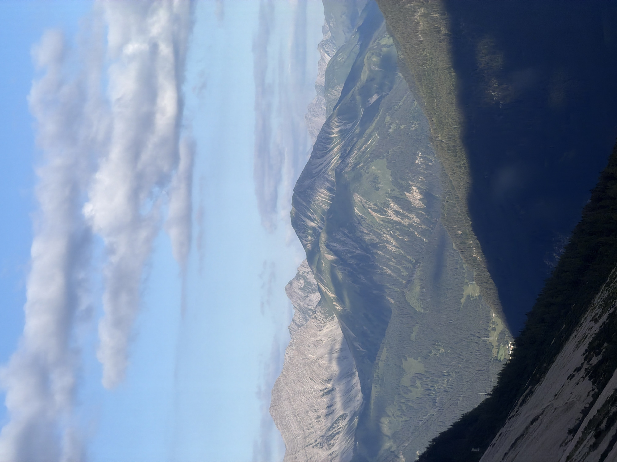 This is a landscape photograph capturing a mountain range under a partly cloudy sky. The foreground shows a steep, rocky slope on the left, transitioning into a dark, densely forested mountainside. A deep shadow is cast across the lower right portion of the image, obscuring details of the terrain. In the mid-ground, layers of green, forested mountains are visible, with some lighter, rocky peaks emerging in the background. The sky is a bright blue, with scattered white and gray clouds. The overall scene depicts a natural mountain environment. The location is identified as Grainau, Germany. There are no people, animals, or specific man-made objects prominently featured that would suggest a particular action or interaction.