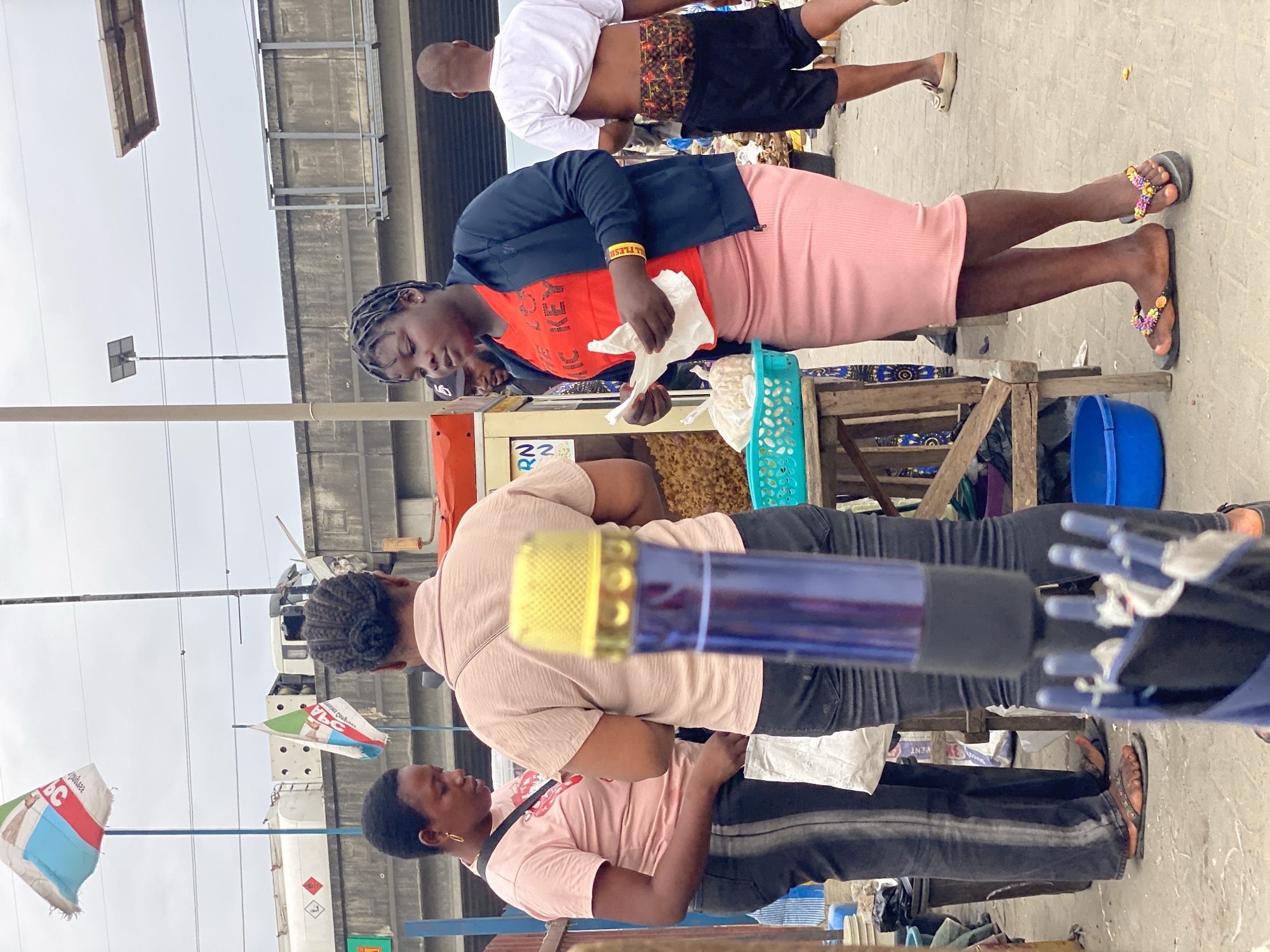 The image captures a street scene, likely in Lagos, Nigeria, due to the presence of election flags affiliated with a Nigerian political party, indicating an election campaign activity. The setting is an open public space with a concrete floor and an elevated structure, possibly an overpass or highway, in the background.
In the foreground, a woman stands behind a popcorn-making machine, filling paper bags with popcorn. She is wearing a black jacket, an orange t-shirt, and a pink skirt, and floral sandals. Behind her, a man with a shaved head is shirtless and wearing patterned shorts.
Further back, two women in pink shirts stand, one wearing black jeans. One is visible to the left and a partial view of the other person is seen. Political flags are on poles in the distance, alongside a vehicle. An umbrella, with its handle in the lower center of the image, partially obscures the view of the two women.