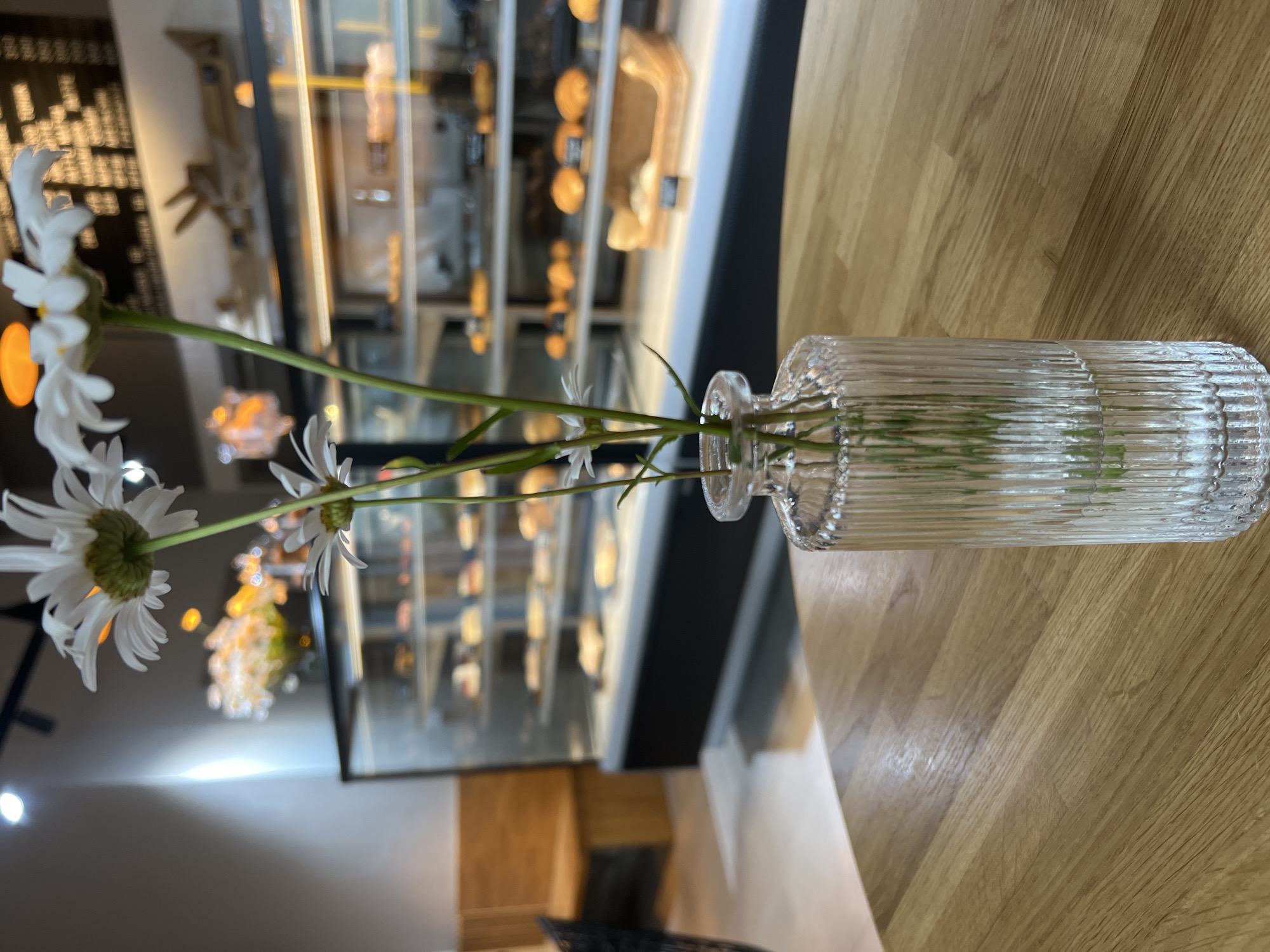 In this image, a clear glass vase holding white daisies stands on a light-colored wooden table. The vase has a ribbed texture. The daisies are white with yellow centers, and their stems are green. In the background, out of focus, is a display case with baked goods. The lighting suggests it might be daytime. The overall scene appears to be indoors, possibly in a cafe or bakery.