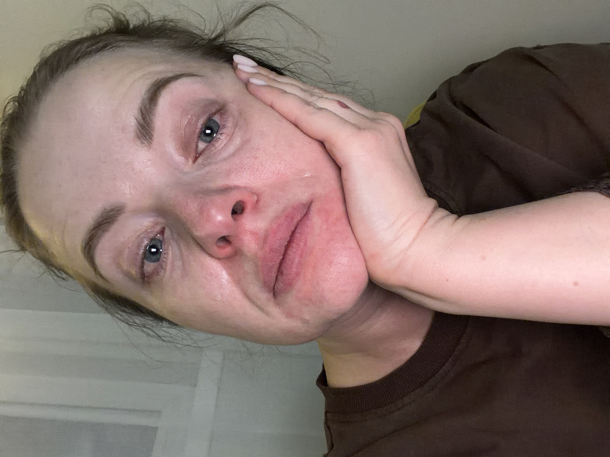 A close-up, low-angle shot depicts a Caucasian woman with visible tear streaks on her face. She appears distressed. Her left hand is positioned against her cheek. She is wearing a brown t-shirt. The background suggests an interior setting. There is no evidence to suggest a location in Moscow, Russia.