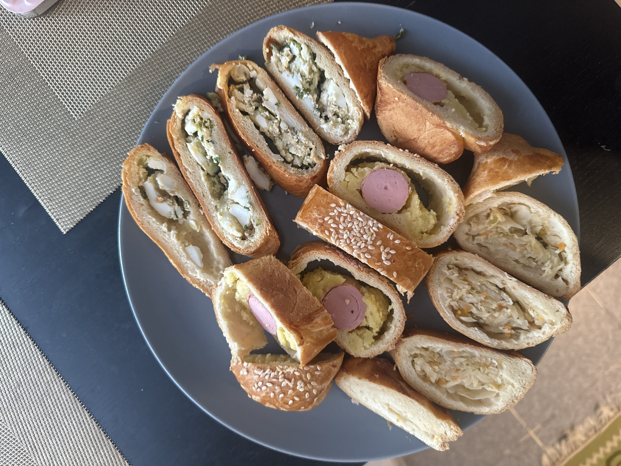 A plate of sliced pastries sits on a table. The pastries are filled with various ingredients, including what appears to be mashed potatoes and sausage, egg and greens, and shredded cabbage with carrots. Some pastries are sprinkled with sesame seeds. The plate is gray and is placed on a dark surface, possibly a table, with a textured placemat to the left. The lighting suggests it is daytime, and the setting appears to be indoors. There are no people visible in the image, and no text is present. The scene is a close-up view of the food, with the pastries being the sole focus. The pastries are varied in their presentation, with some cut lengthwise and others crosswise. The colors are warm, with the golden brown of the pastry contrasting with the lighter fillings. The overall impression is one of a home-cooked meal or a prepared snack. The location context of Krasnoyarsk, Russia, does not provide any specific visual cues within the image itself.