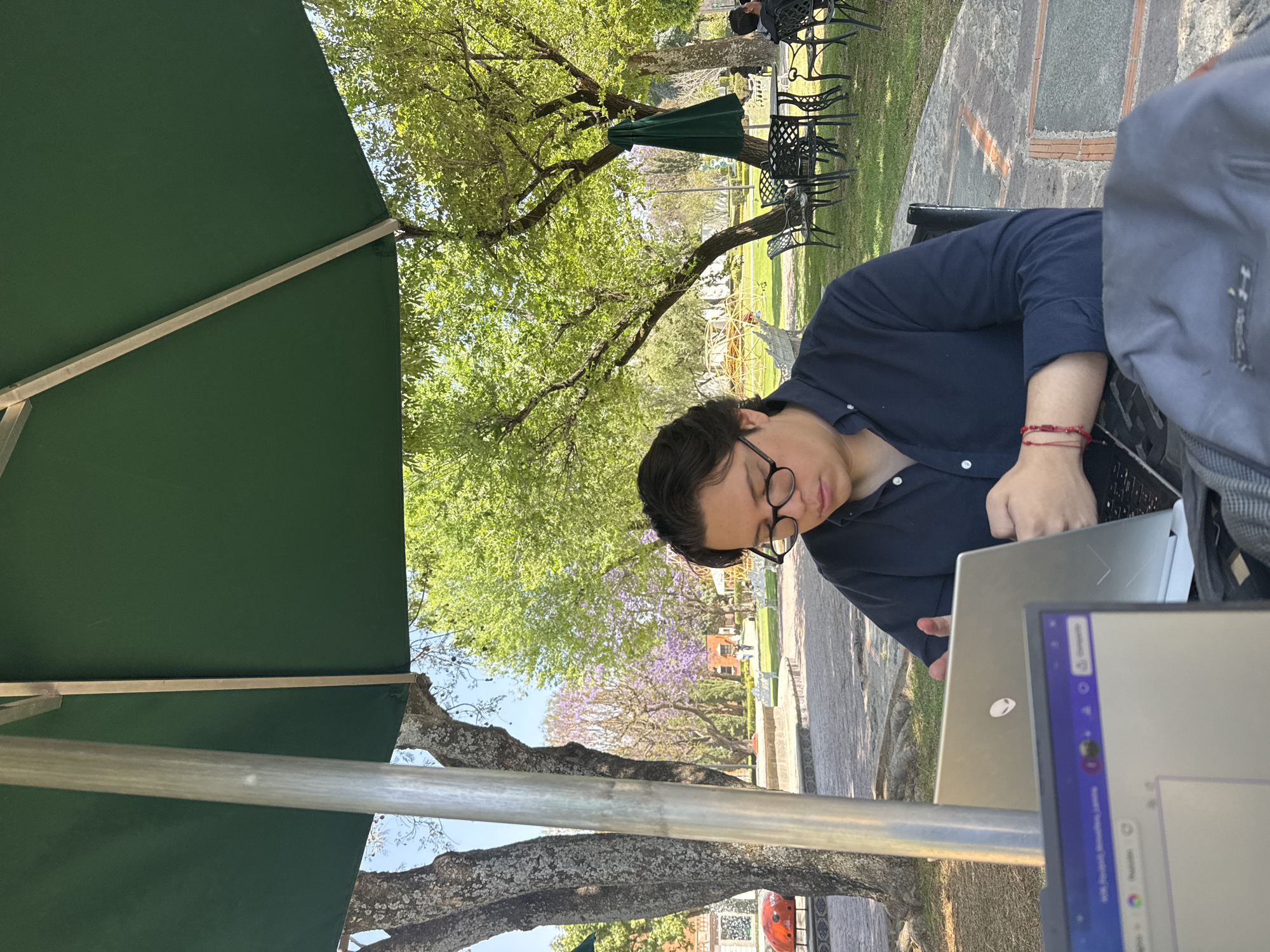 The image depicts an outdoor scene in a public park or plaza in San Andrés Cholula, Mexico, under a clear, sunny sky. A light-skinned male with dark hair and glasses, wearing a dark blue long-sleeved shirt and two red string bracelets on his right wrist, is seated at a dark table. He is focused on a silver-colored laptop, identifiable by an Alienware logo, positioned in front of him. A second laptop with visible browser tabs is partially obscured in the immediate foreground. A grey backpack rests near his right arm. A large, dark green rectangular umbrella provides shade directly above the subject.
The immediate surroundings feature a paved stone surface transitioning into grassy areas. Mature trees with both bright green leaves and purple blossoms (suggestive of Jacaranda) are abundant throughout the park. In the midground, multiple dark metal tables and chairs are visible on the grass, with one set positioned under a folded green umbrella. White park benches are interspersed along pathways. A large red, spherical sculpture, resembling a ladybug, is situated on a stone fountain or planter in the left background. Distant buildings with reddish roofs are visible. A second, partially obscured individual is seated at a table in the far right background, partially concealed by a tree and another umbrella.