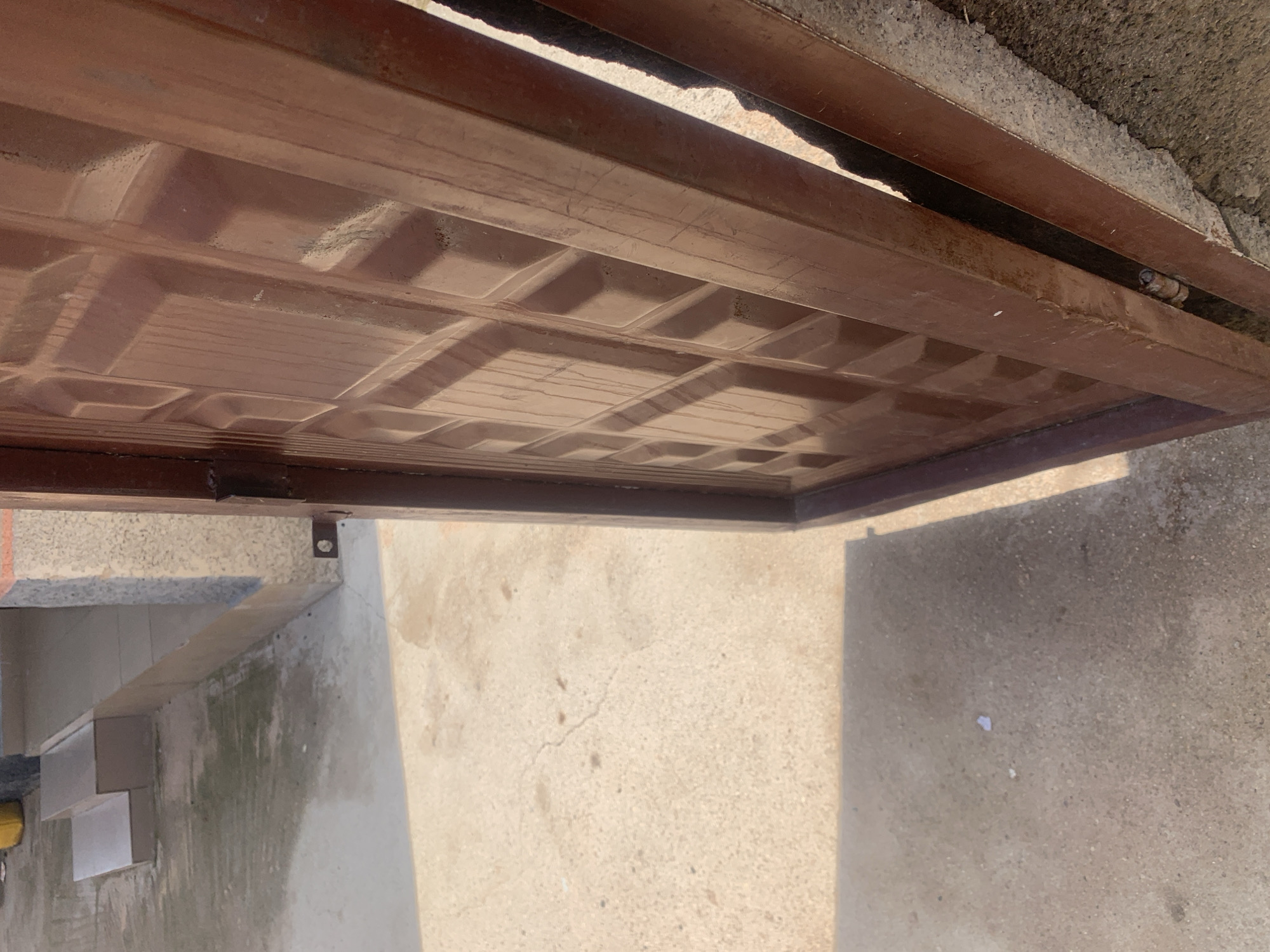 The image depicts an outdoor scene, likely in Jauro Yinu, Nigeria. In the foreground, a partially opened brown metal gate with a geometric pattern is visible. The gate's frame appears to be made of metal and shows signs of rust. To the left of the gate, a concrete ground surface is visible, with some dark stains and cracks. Further left, there is a set of steps leading up to a building. The steps and the adjacent wall are tiled. In the background, a yellow object, possibly a container, is partially visible. The lighting suggests it is daytime. The scene appears to be an entranceway or a pathway leading to a building. There are no people or animals clearly depicted in the image.