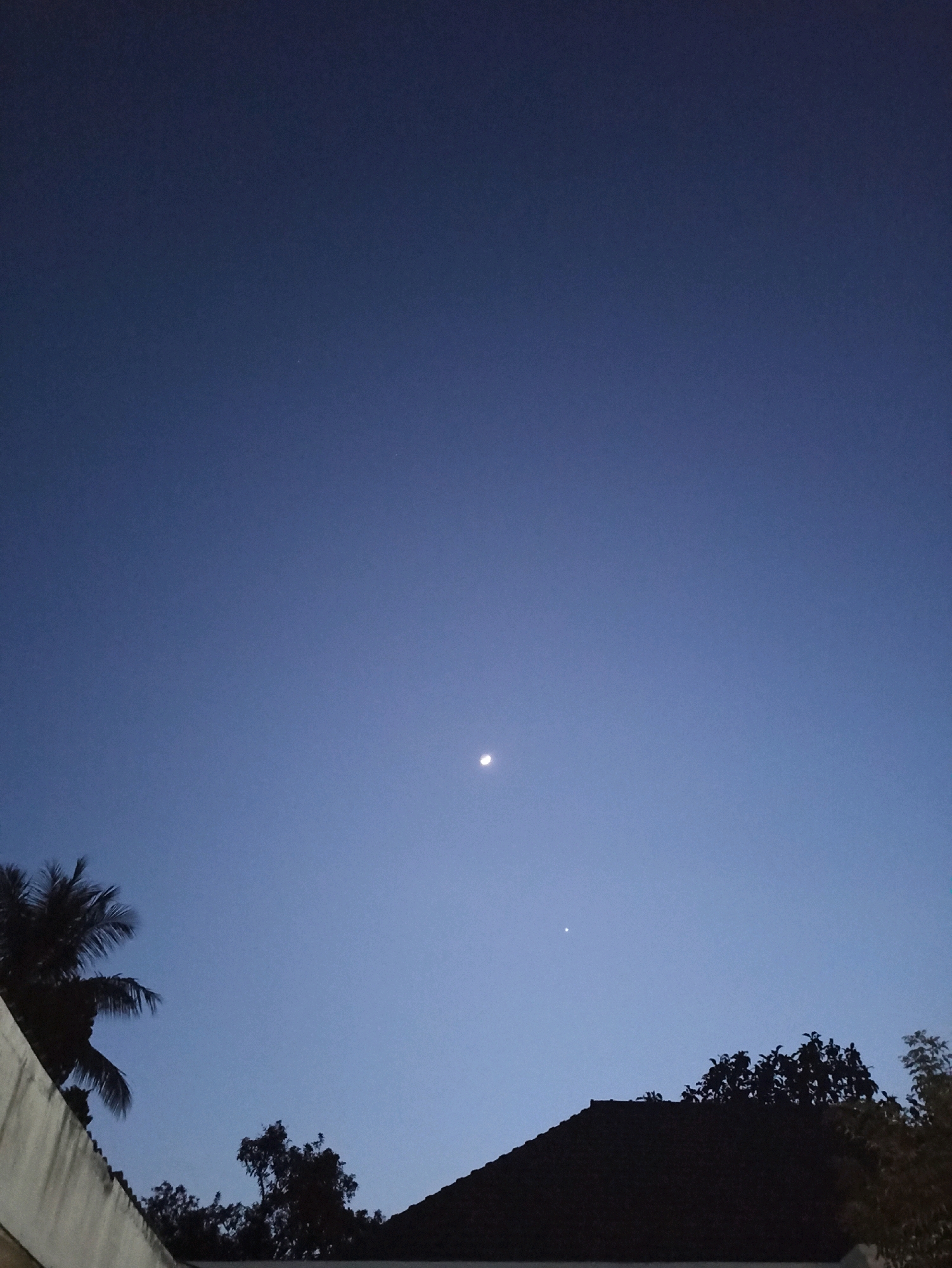 The image depicts a twilight sky above a residential area. In the center of the frame, a sliver of the moon is visible, accompanied by a bright point of light, likely a planet. Silhouetted against the sky are the rooftops of houses, some trees, and a portion of a building with a textured wall in the foreground. A palm tree with its fronds is visible on the left side of the image. The sky is a deep blue, gradually lightening towards the horizon. The scene appears to be set in Brambang Darusalam, Indonesia, given the tropical vegetation. There are no people visible in the image.