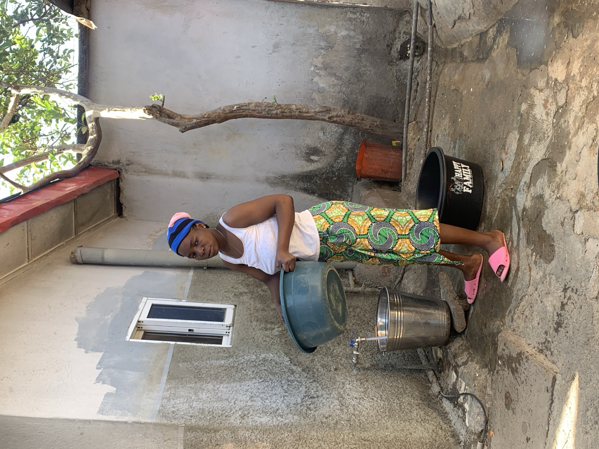 A young woman is captured in an outdoor utility area or courtyard, likely in a residential setting in Jos, Nigeria, during bright daylight. She stands facing forward, looking towards the camera with a neutral expression.
The woman wears a white tank top, a brightly colored, patterned skirt featuring green, orange, and yellow concentric circle designs, and pink slide sandals. Her head is covered with a blue and pink knitted cap. She is holding a large, green plastic basin with both hands, positioned near a metal bucket. A small, silver-colored tap is visible on the concrete ground next to the metal bucket, suggesting she is engaged in an activity related to water collection or usage. The area around the tap and buckets appears wet.
The setting is characterized by concrete walls and ground, showing signs of wear and discoloration. To the left of the woman, a small, horizontal window with a white frame is set into the wall. Above the woman, a sturdy, gnarled wooden branch extends across the frame, with some green foliage visible on the far left, suggesting natural elements within the enclosed space. Further back and to the right, a black plastic bucket is visible, bearing the white text "HAPPY FAMILY". An orange-red plastic planter is also seen in the background.