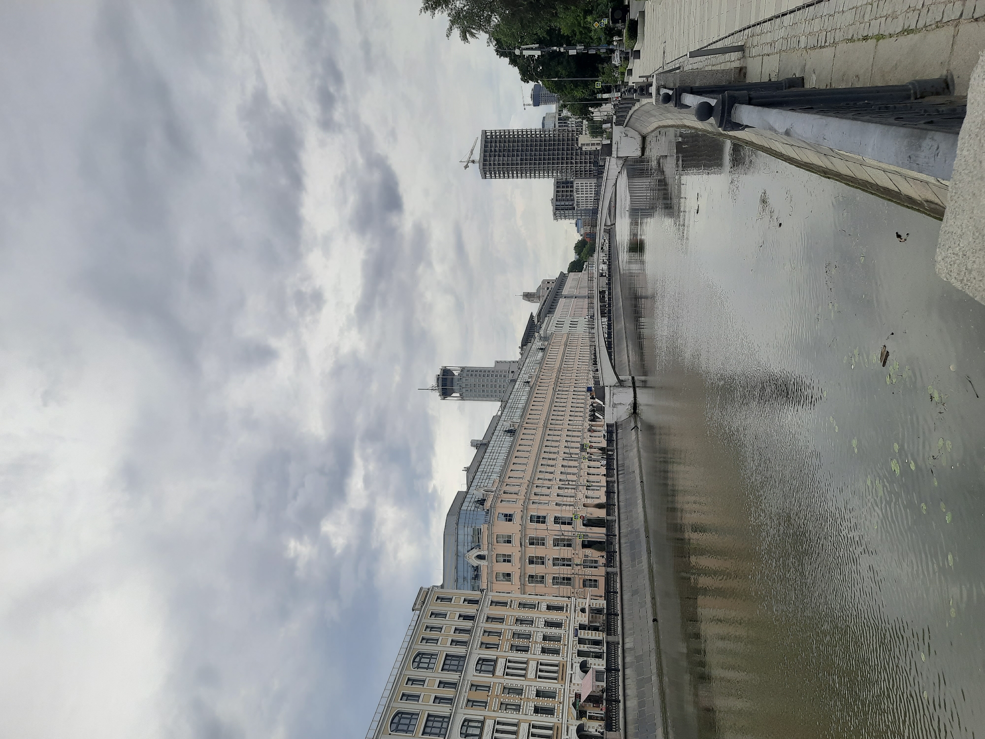 The image depicts a wide river flowing through a city under a cloudy sky. Along the riverbank, a paved walkway is visible, bordered by a black metal railing. Several multi-story buildings line the opposite bank, some appearing to be historical architecture with many windows, while others are more modern with glass facades. In the distance, a tall skyscraper with a distinctive spire can be seen, along with other modern buildings, one of which appears to be under construction with a crane on top. A white bridge spans the river in the middle ground. The water has a greenish-brown hue and small lily pads are scattered on its surface. The overall impression is of an urban waterfront scene on an overcast day.