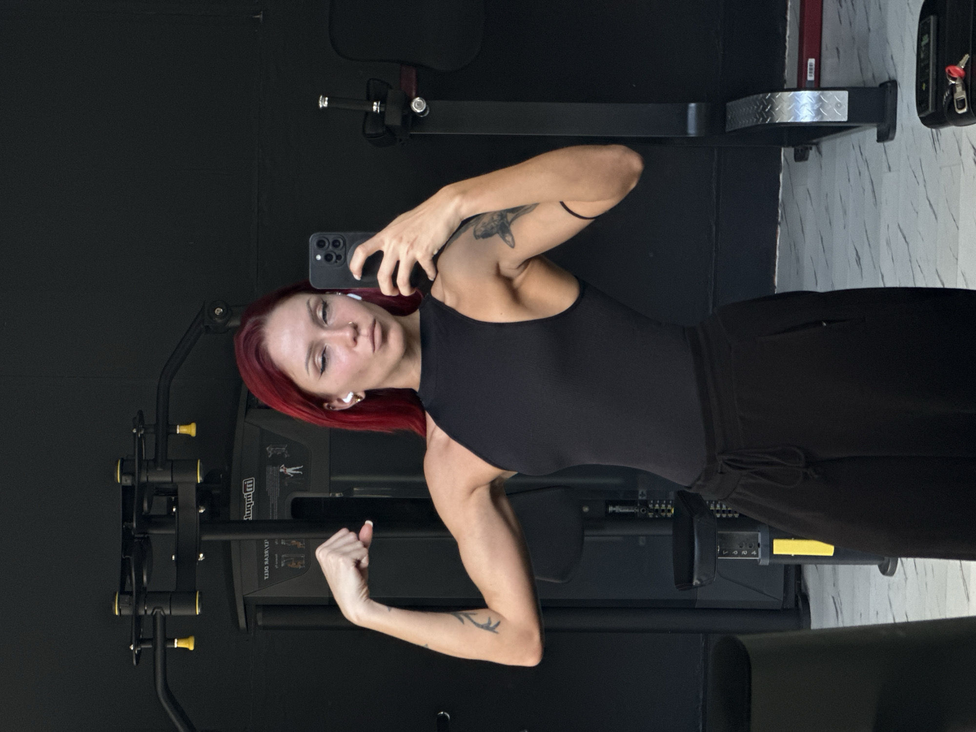 A woman with red hair is taking a selfie in a gym. She is wearing a black tank top and dark pants, and she is flexing her left arm. She is holding a smartphone in her right hand. In the background, there are various pieces of gym equipment, including a black weight machine and another metallic one behind her. The location appears to be a gym in Tibubeneng, Indonesia.