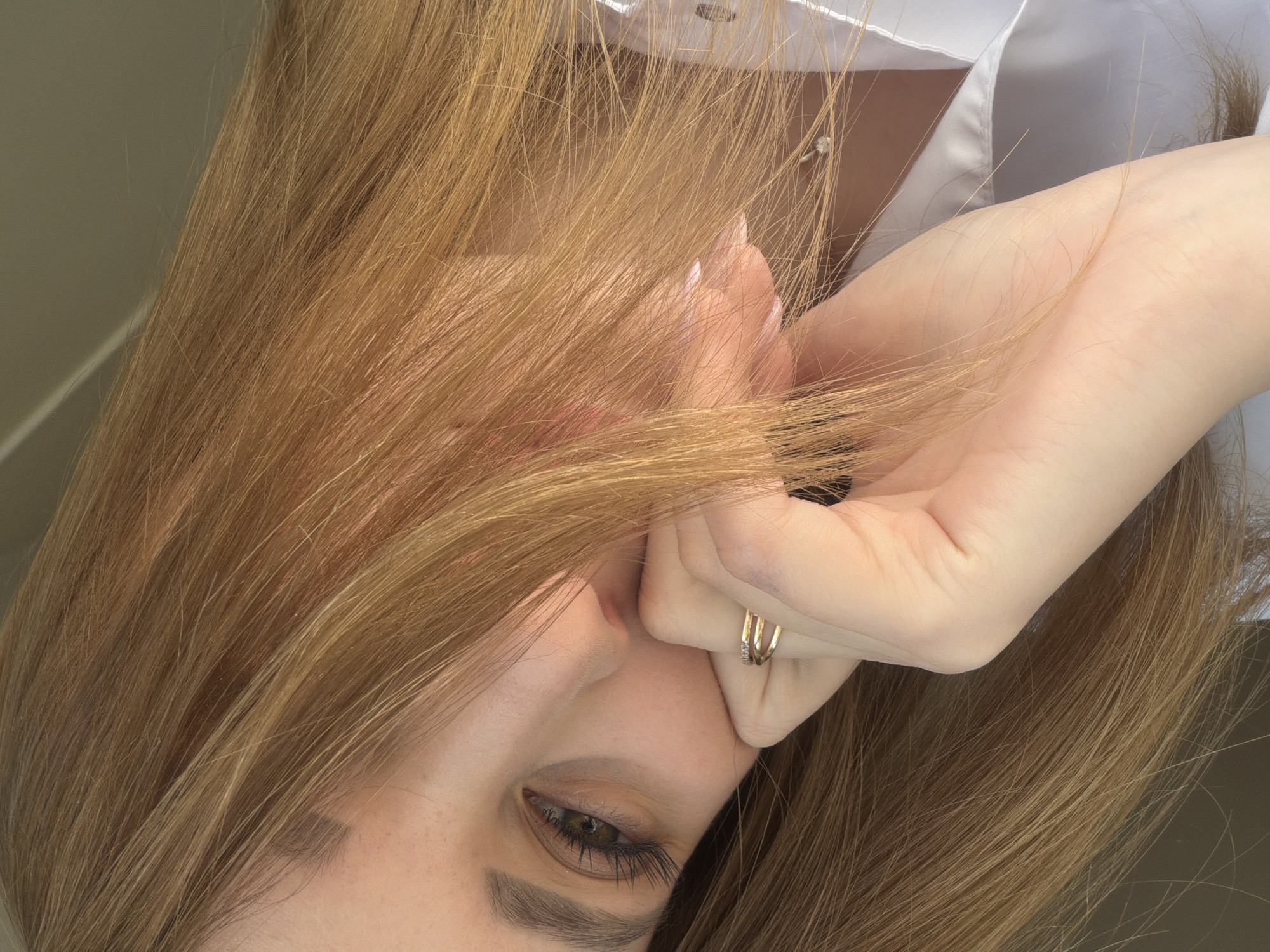 A selfie-style close-up photograph shows a woman with long, light brown hair partially obscuring her face. Her left eye is visible and brown. Her hand, adorned with a gold ring featuring a row of small stones, is raised to her forehead, with strands of hair caught between her fingers. She is wearing a white shirt. There is no identifiable information to confirm the location is Chaikovsky, Russia.