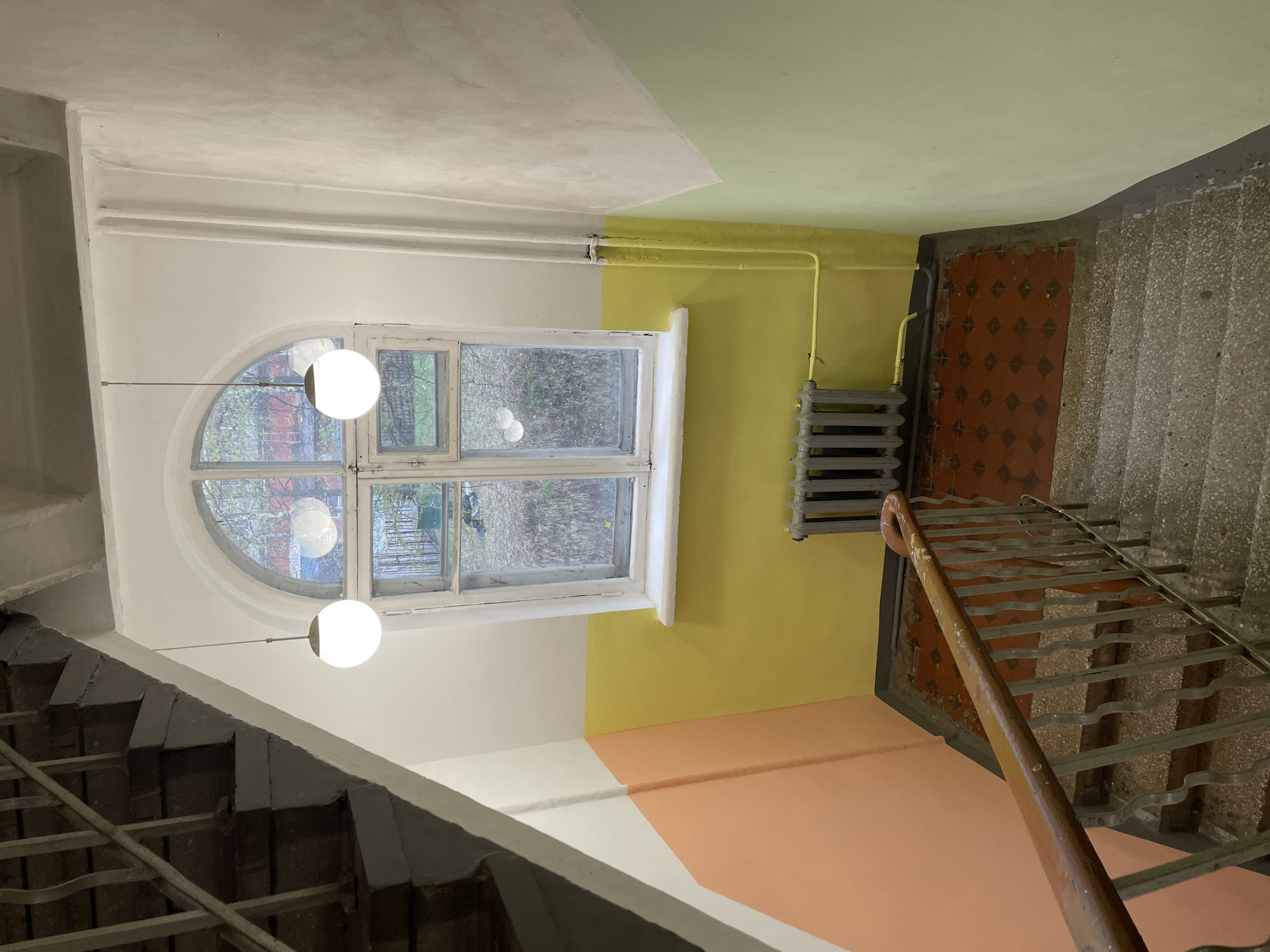 This image presents an interior view of a stairwell and landing within a building, identified as being in Khimki, Russia. No people are present, and no specific activity is depicted, making it a static scene.
The focal point is a large, multi-paned window with a distinct rounded arch at its top, framed in white. Through its clear glass, bare tree branches and hints of other buildings are visible, suggesting an exterior view during an overcast day or a cooler season. Two modern, spherical white pendant lights hang at varying heights near the window, providing artificial illumination.
The walls are painted in a striking, angular color-block pattern. The wall immediately surrounding the window is white. To its right, the wall transitions to a vibrant yellow section. Above the yellow wall, the ceiling is painted in a light green or lime hue, which sharply contrasts with the white ceiling section over the white wall. Below the yellow wall, a lower wall section and part of the landing area are painted in a soft peach or light orange color, meeting another section of white. A gray, cast-iron radiator is mounted on the yellow wall, with yellow pipes running above it.
To the right, a robust staircase descends, featuring dark gray, concrete or terrazzo-like steps. A sturdy metal railing with a worn, warm-toned wooden handrail provides support. A section of the floor at the base of the stairs is adorned with reddish-brown tiles, featuring a repeating dark diamond pattern.
The overall impression is of an older architectural space that has undergone a modern renovation, blending traditional structural elements with contemporary lighting and a bold, colorful painting scheme. There is no visible text in the image.