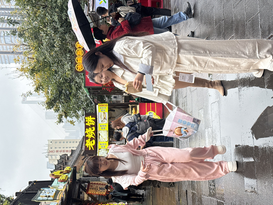 This image depicts an overcast day on a wet, paved pedestrian street in Yuzhong District, Chongqing, China. The scene features three young adult females in the foreground, with numerous other individuals, vendors, and traditional-style storefronts visible in the mid-ground and background. Distant high-rise buildings and a cable-stayed bridge are visible under a grey sky.
In the left foreground, a female with long dark hair, wearing a light pink hooded tracksuit and white platform sneakers, walks left-to-right. She holds a light pink shopping bag with a blue cartoon keychain in her left hand and a white smartphone in her right, looking slightly downwards. To her right, a second female with long dark hair stands, smiling faintly. She wears an off-white short-sleeved top and tan knee-high boots, holding a small brown paper bag, likely containing food. Adjacent to her, a third female with long dark hair, wearing an off-white jacket with striped cuffs, matching wide-leg pants, and white sneakers, stands facing downwards, focused on a white smartphone held in both hands. A white tag hangs from her jacket.
The street surface is dark grey, wet, and reflects light from puddles. To the left, traditional Chinese architecture with dark roofs and elaborate signage, featuring Chinese characters, lines the street. One prominent yellow sign in the mid-ground clearly displays "老烧饼" (Lǎo Shāobǐng), indicating a food stall. Further right, trees with green and some yellowing foliage line the street. In the mid-ground, various individuals are present, including a person in a red jacket holding a black umbrella to the right. The background features modern high-rise buildings under construction or completed, and a large cable-stayed bridge spanning a body of water, all partially obscured by the overcast conditions.