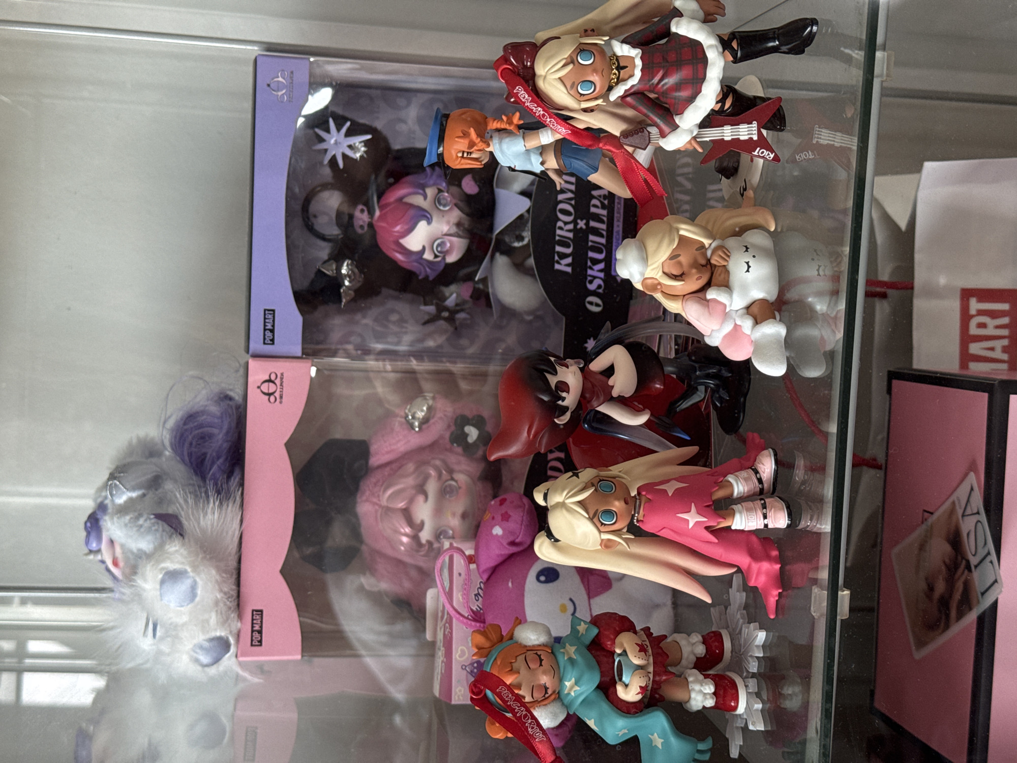 Here's a detailed description of the image:
**Setting:** The image appears to be taken inside a display cabinet or shelf. There are various figurines and boxes, suggesting a collection of some kind.
**Objects:**
* A white and gray plush toy with purple accents, possibly a character from a fantasy or anime series.
* Several boxes from POP MART, which is a company that produces collectible toys and figurines.
* Several collectible toys are not in boxes.
* There's a card with the name "LISA" written on it.
* The text "PEACHORIOT" is visible, likely as part of the branding on one of the items.
* The text "KUROMI X SKULLPANDA" is visible, possibly part of the name of a figurine or collection of figurines.
* One of the figurines is holding a red guitar.
* Text "RIOT" is visible next to the red guitar.
**Composition:** The figurines and boxes are arranged in a somewhat cluttered manner within the cabinet, creating a sense of depth.
Based on the information available to me, I cannot determine the location in the city of บ้านกาญจน์กนก 23, Thailand.