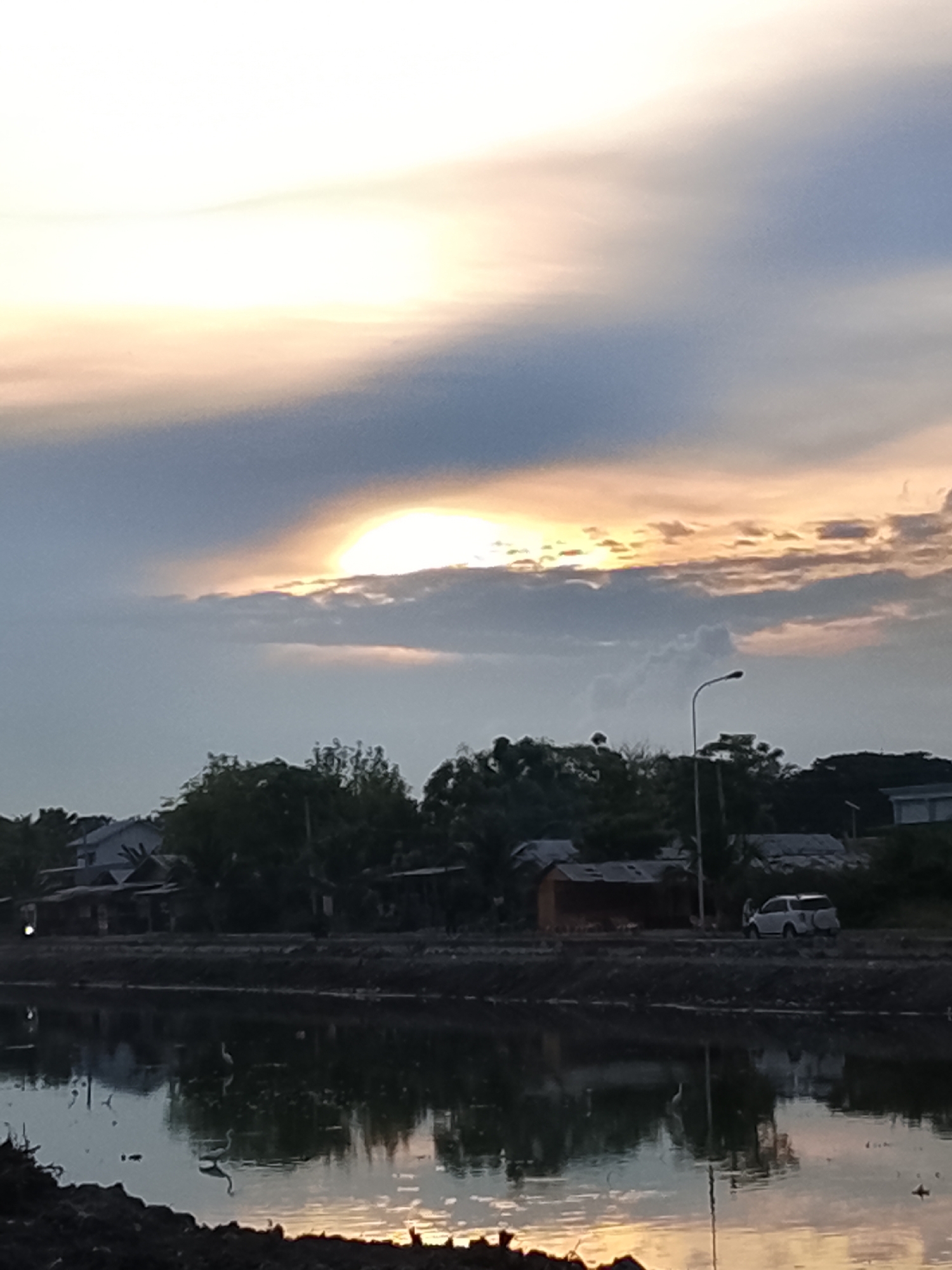 The image depicts a sunset scene in Lhokseumawe, Indonesia, featuring a body of water in the foreground reflecting the sky. The sky is partly cloudy with the sun peeking through the clouds, casting a warm, golden light. In the middle ground, there is a line of trees and some low-lying buildings. A white SUV is parked on a dirt road adjacent to the water. Several birds, possibly egrets, are visible in the water and on the shore. The overall impression is a serene, natural landscape within an urban or semi-urban environment.