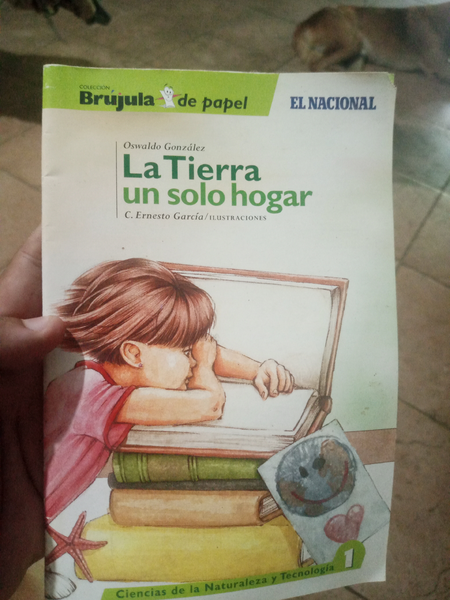The image displays the cover of a book titled "La Tierra un solo hogar" by Oswaldo González, with illustrations by C. Ernesto García. The cover features a watercolor illustration of a child resting their head on a stack of books. A drawing of the Earth with a smiley face and a heart is placed near the books. The book is part of the "Brujula de papel" collection and published by EL NACIONAL. The bottom of the cover has the text "Ciencias de la Naturaleza y Tecnología". The image is taken from a slightly elevated angle, showing a hand holding the book. The background is blurry, but appears to be an indoor setting with a tiled floor and a hint of an animal in the upper portion. The lighting suggests it's daytime. The location is identified as Santa Ana, Venezuela.