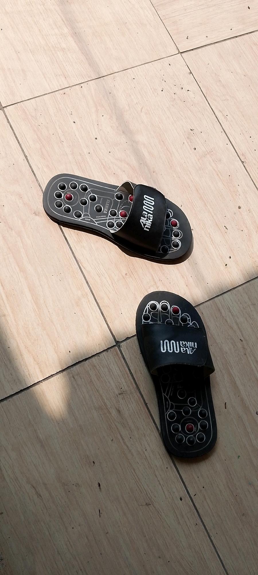 Two black reflexology sandals are placed on a tiled floor, which appears to be outdoors due to the lighting and shadows. The tiles are a light beige color with visible grout lines. The sandals have a black strap with a white logo that reads "Ala Nika 1000". The soles of the sandals are black and feature a pattern of circular bumps, some red and some black, arranged to correspond with reflexology points on the feet. Text labels are visible on the sole of the sandal in the upper left of the image, indicating areas like "ANUS" and "PANCREAS". The sandals are positioned with the left sandal slightly above and to the left of the right sandal. The overall scene is static and focuses on the objects. The location is identified as Satriamekar, Indonesia.