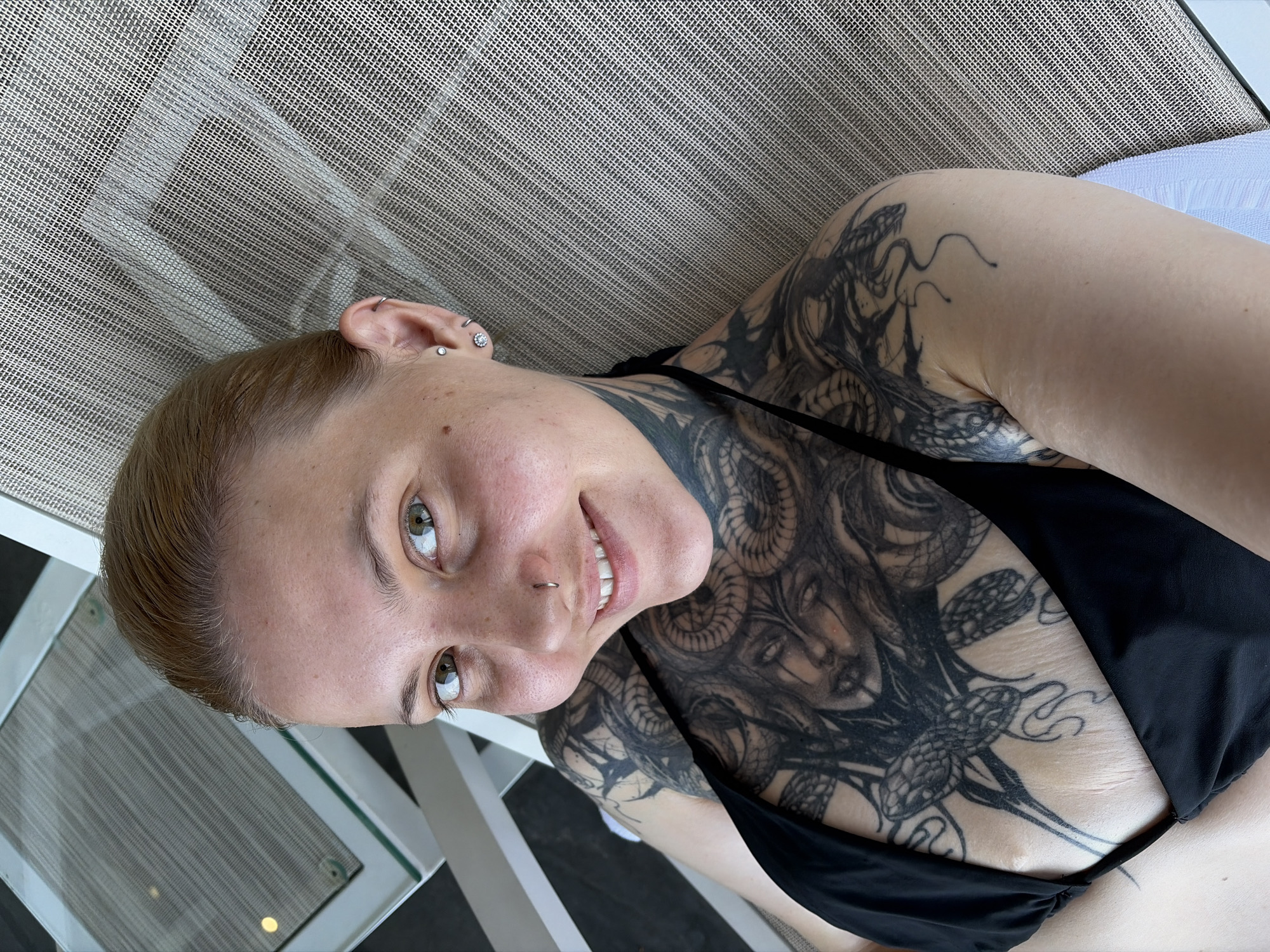 A woman with light brown hair and striking green-hazel eyes is captured in a close-up, smiling portrait. Her head is tilted, giving the image a rotated appearance. She has a small septum piercing and multiple ear piercings, including a stud in her tragus and a jeweled stud in her lobe. Her complexion shows light freckles.
A significant portion of her chest, neck, and left shoulder is covered with intricate black-ink tattoos. The design prominently features a stylized face, possibly Medusa, with numerous intertwining snakes extending up her neck and over her shoulder and arm. She is wearing a black bikini top.
The woman is reclining on light-colored outdoor furniture, characterized by a woven, mesh-like texture, suggesting a lounge chair or daybed. A white frame and a reflective surface, possibly a table, are partially visible in the background. The scene is brightly lit by natural light, indicating daytime. She appears relaxed and content, consistent with enjoying leisure time in an outdoor setting such as a resort or poolside area in Đà Nẵng, Vietnam. No text is visible in the image.