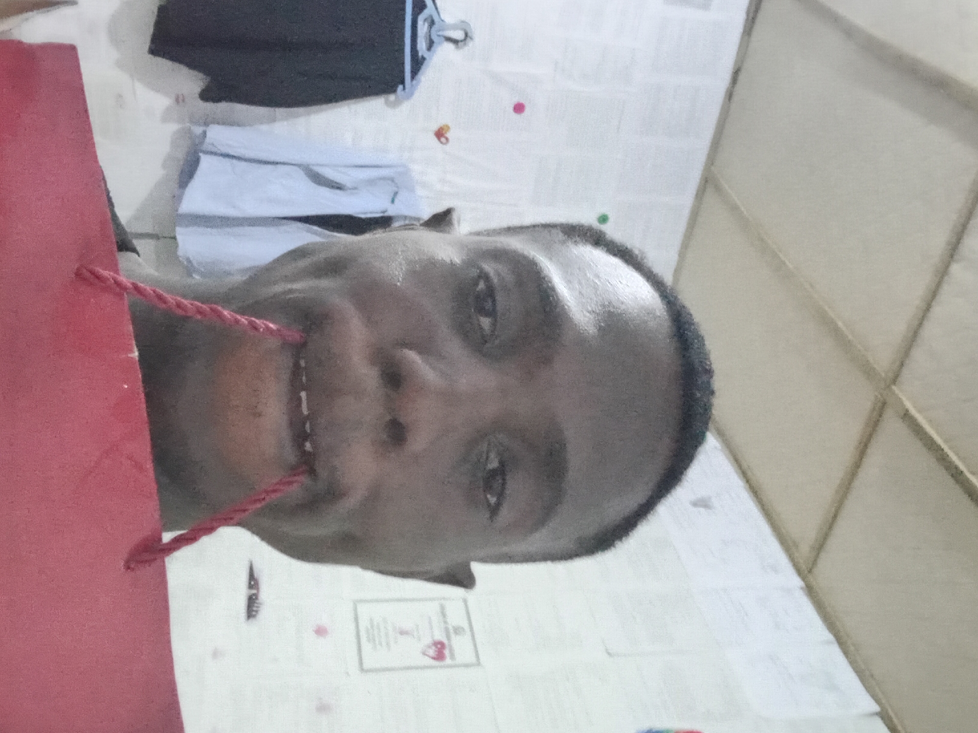 A medium shot shows a person from the chest up with one red bag strap placed in their mouth. The individual appears to be of African descent, with dark hair and eyes. They are indoors. Behind them are clothes and what appear to be walls or doors with a grid pattern, possibly made of light wood. The location is presumed to be Akwete, Nigeria.