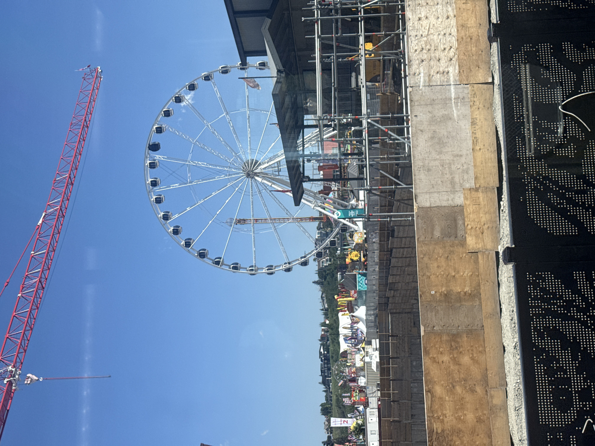 The image captures a sunny day with a clear blue sky, showcasing a construction site in Calgary, Canada. Dominating the scene is a large Ferris wheel, a temporary structure likely part of a fair or event. Adjacent to the Ferris wheel, a red construction crane extends from the upper left corner, with its boom angled across the sky.
The foreground reveals the ongoing construction with scaffolding surrounding a low-rise building. Wooden planks and concrete walls form barriers around the excavation site. In the middle ground, various stalls and tents associated with an event are visible, creating a festive atmosphere beneath the Ferris wheel. The background features a gentle slope covered with trees and residential buildings, suggesting a suburban or urban fringe setting. The overall impression is one of a dynamic urban landscape where recreation and development coexist.