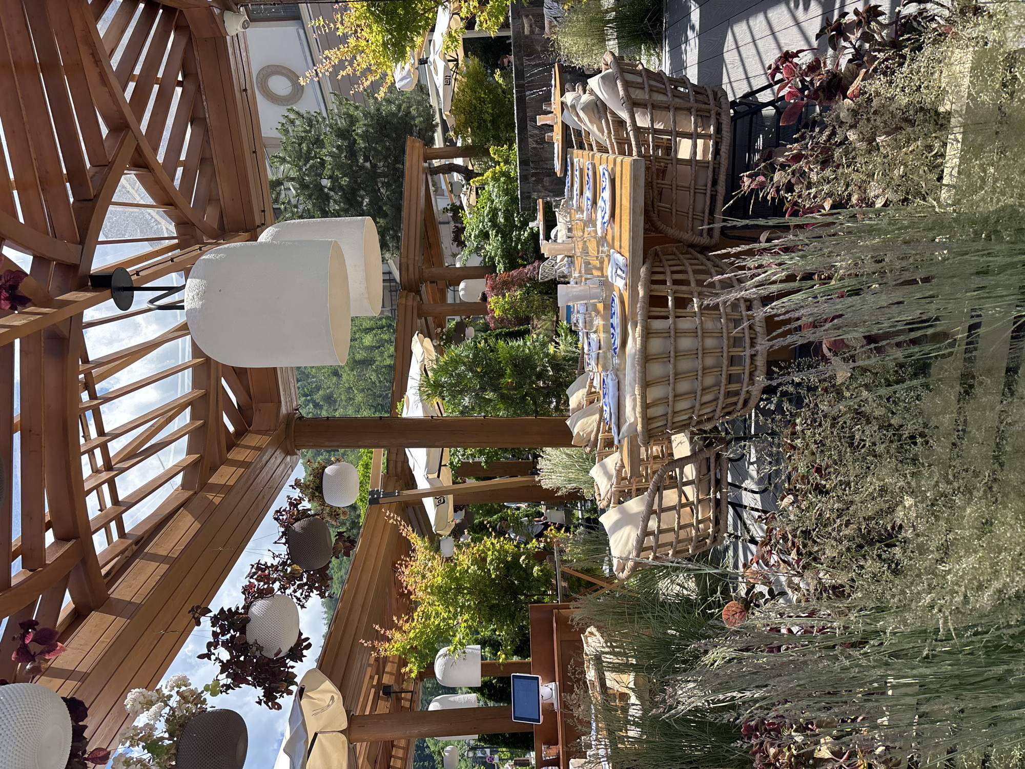 This is a medium shot, taken outdoors during the daytime, of a restaurant seating area with a pergola roof in Estosadok, Russia. The pergola is constructed of natural wood and has a slatted roof, allowing sunlight to filter through. Hanging from the pergola are large, white, bell-shaped lamps and several hanging planters with greenery and flowers.
In the foreground, there is a lush arrangement of tall, feathery grasses and dark red foliage. Behind this, a large wooden table is set for dining with blue and white patterned plates, glassware, and white napkins. Woven wicker chairs with beige cushions surround the table.
Further back, more tables and chairs are visible under additional pergolas and umbrellas, suggesting a spacious outdoor dining facility. A person is visible walking in the background. Trees and foliage create a green backdrop, and a building with a circular decorative element can be seen in the distance. The overall impression is one of a well-appointed and inviting outdoor restaurant space.