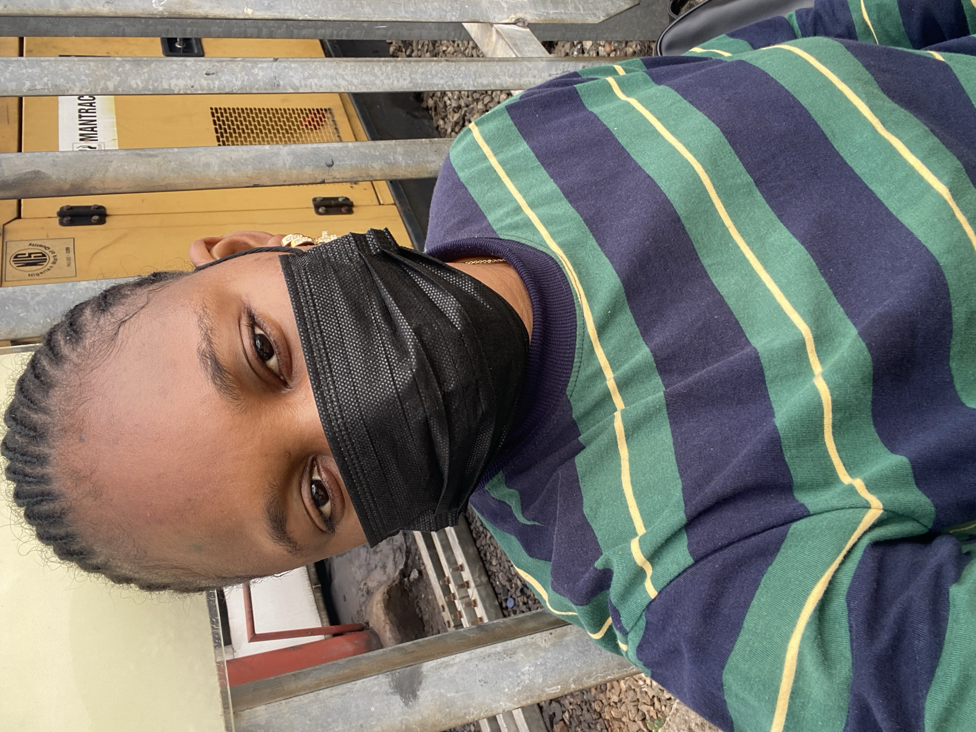 The media file shows a close-up, vertically oriented, selfie-style image of a young Black person wearing a black disposable face mask and a navy blue, green, and yellow striped long-sleeved shirt. Their hair is braided. They are wearing gold jewelry on their ear and a ring on one finger. The person is looking towards the camera, with their eyes visible above the mask.
In the background, a yellow industrial generator is partially visible, with the word "MANTRAC" and a logo that appears to read "NIGERIA" printed on it. There are also metal railings and gravel. The scene suggests an outdoor setting, possibly near a construction site or industrial area. The lighting indicates it could be daytime. The overall emotion conveyed is neutral, with the mask obscuring much of the person's facial expression. The context provided places the location in Keffi, Nigeria.