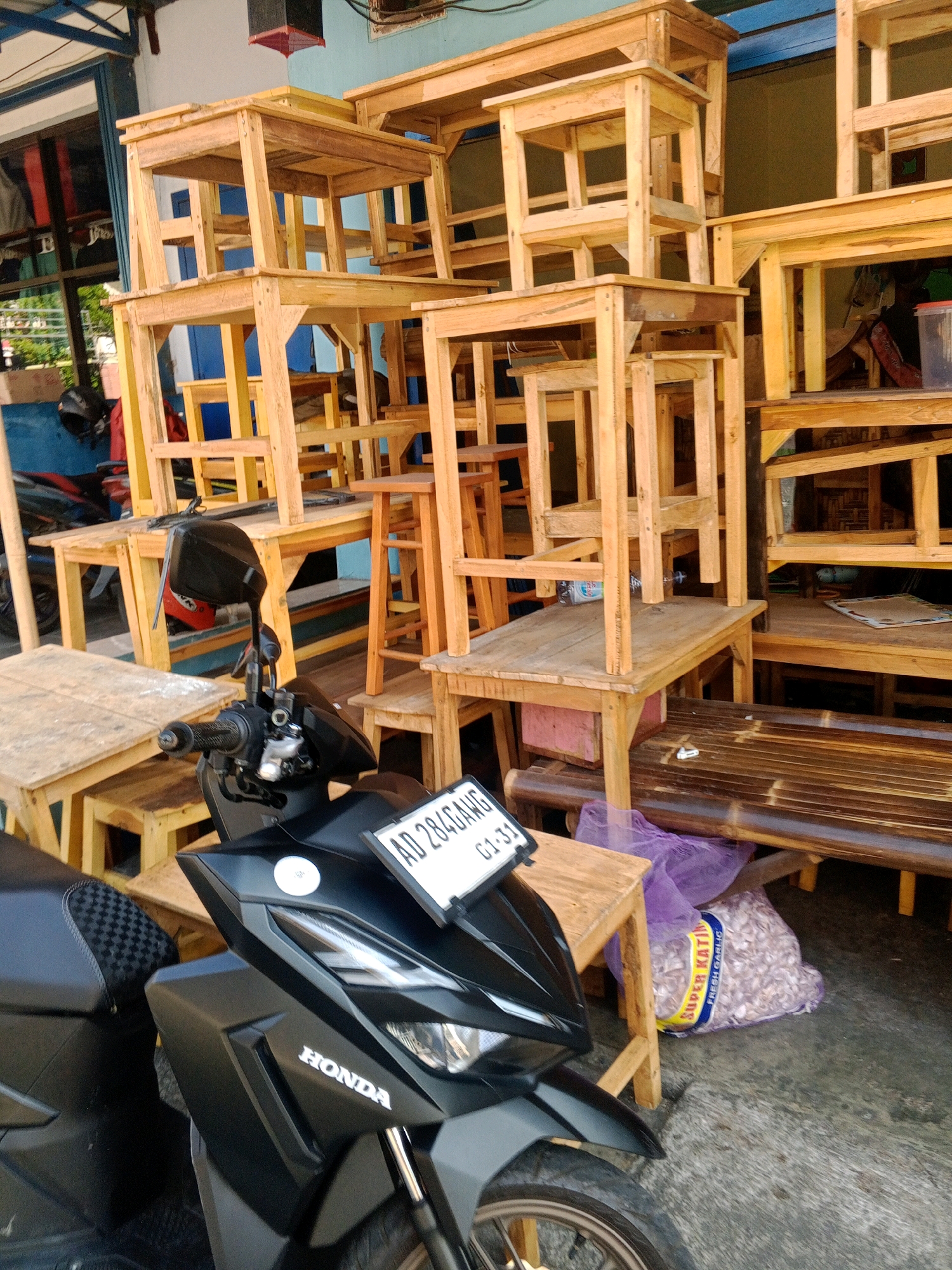 In a street-side setting in Wonogiri, Indonesia, numerous simple wooden tables and chairs are displayed for sale. The furniture is constructed from light-colored wood, with a stacked and somewhat haphazard arrangement. In the foreground, a black Honda scooter is parked. The scooter has a license plate with the characters "AD 284GANG" visible. A large, clear plastic bag filled with what appears to be garlic is situated near the scooter and under one of the wooden tables. The background shows a building with a blue facade and some signage, suggesting a commercial area. The lighting indicates it is daytime, with sunlight casting shadows.