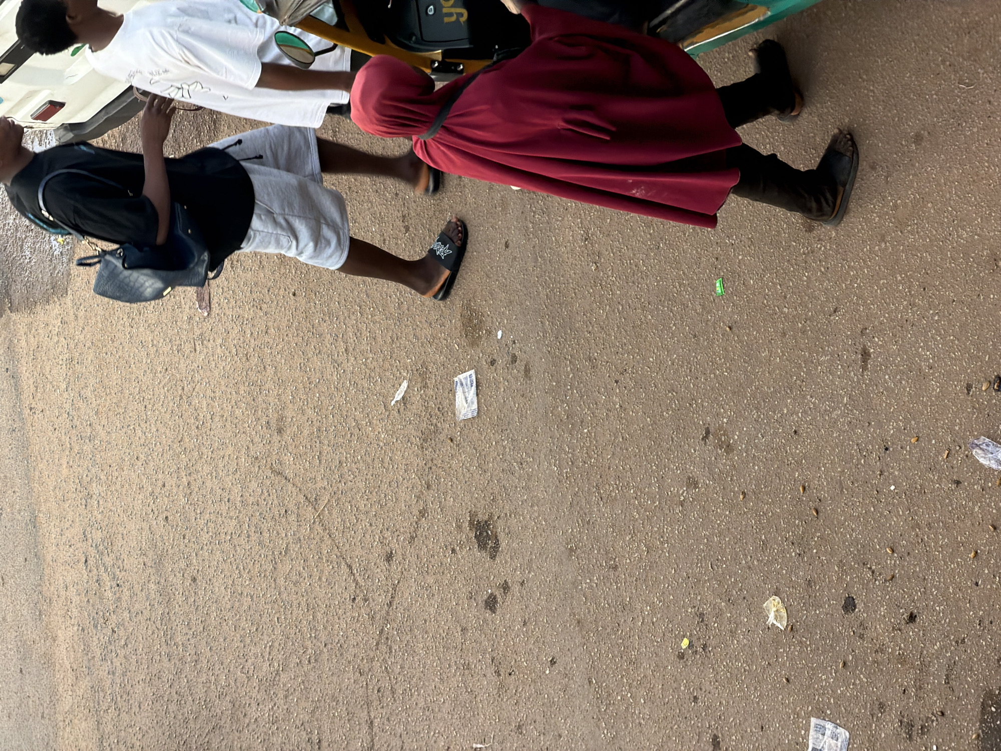 In Bukuru, Nigeria, an outdoor scene unfolds on a dusty, asphalt-like surface, potentially a road or street. Three individuals are visible. In the foreground, a person wearing a crimson-colored hooded garment and dark pants with open-toed sandals is partially visible, interacting with a yellow and green vehicle. Behind them and to the left, another person is seen from the waist down, wearing light gray shorts, a black t-shirt, and black slides with white lettering. This individual carries a dark backpack. To the right of this person and slightly further back, another individual is seen wearing a white t-shirt. The ground is littered with various small debris, including what appears to be wrappers or paper scraps and some small, dark spots. The overall impression is one of a casual, everyday moment on a street in a Nigerian city.