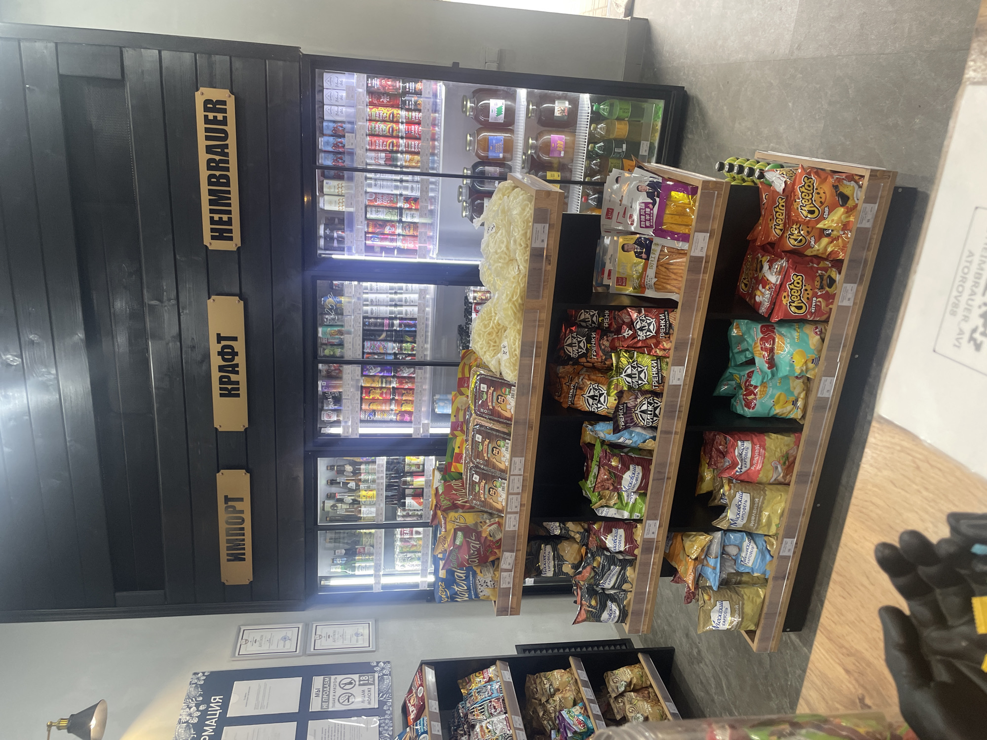 The image shows the interior of a small store or market. In the background, there are three large glass-door refrigerators filled with beverages. Above the refrigerators, signs are mounted on a dark wooden wall. The signs read "ИМПОРТ" (Import), "КРАФТ" (Craft), and "HEIMBRAUER".
In the foreground, shelves are stocked with various food items, primarily snacks and chips. There are bags of potato chips, Cheetos, and other packaged snacks. Some of the bags have Russian text on them, indicating brands like "Московский картофель" (Moscow Potatoes) and "ФЛУШКА" (Flushka). There are also jars of preserves or drinks in the refrigerator.
To the left of the refrigerators, a black shelf unit displays more packaged snacks. A blue informational poster is mounted on the wall to the left, with text including "Мы не продаем табак и алкоголь" (We do not sell tobacco and alcohol).
A wooden counter is visible in the lower part of the image, with a hand wearing a black glove resting on it. A partially visible document with text like "ATOROVSS HEIMBRAUER AVT" is on the counter.
The overall environment suggests a retail space selling imported goods, craft products, and snacks. The presence of the "HEIMBRAUER" sign and the mention of it on the document suggest a connection to the city of Zhukovo, Russia, where this brand may have a presence.