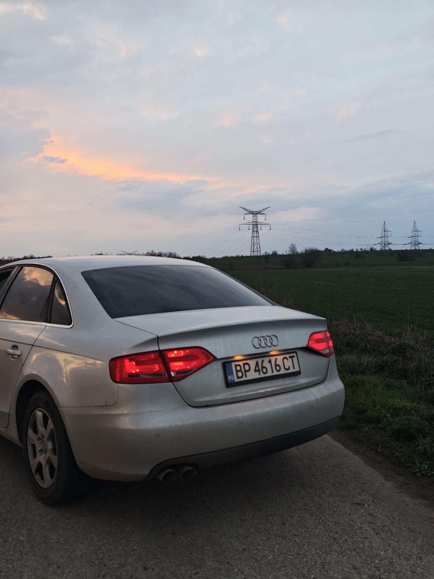 A silver Audi sedan, likely an A4, is depicted from its rear three-quarter angle, parked stationary on the right side of a single-lane paved road. Its taillights are illuminated red. The Bulgarian license plate reads "BP 4616 CT." The car's rear bumper features visible parking sensors and dual exhaust pipes on the left. The rear window is tinted. To the right and extending into the background are expansive green fields, with a sparse treeline visible further back. Multiple large metal lattice transmission towers (power pylons) carrying power lines span the distant landscape; the most prominent pylon is situated towards the left-center background. The sky above is largely overcast with an orange and pink glow visible on the horizon towards the left, indicating dusk or sunset. No individuals are present in the scene. The setting is a rural or semi-rural area near Gorni Tsibar, Bulgaria.