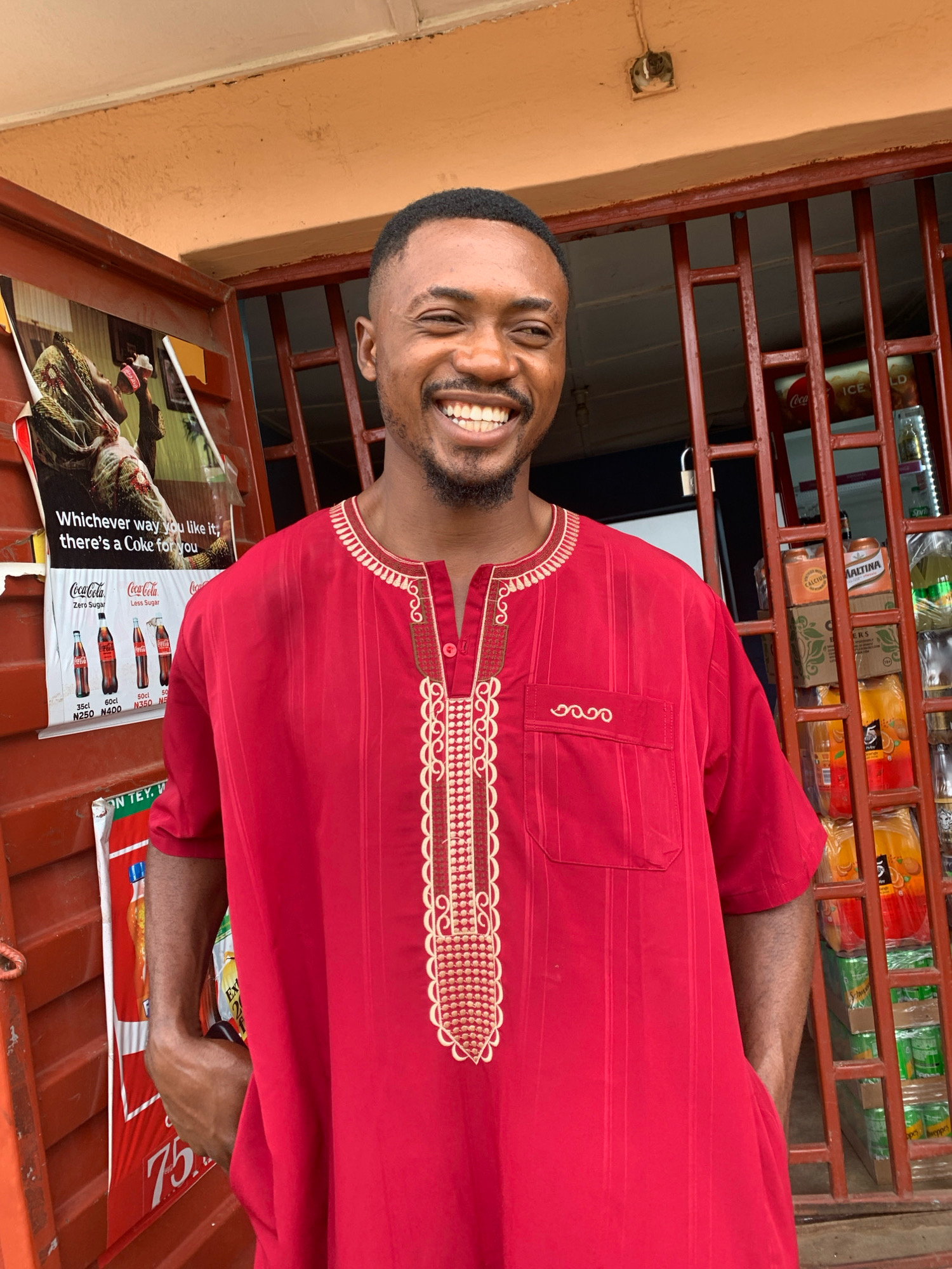 A Black man with short hair and a beard smiles broadly at the camera, his head tilted slightly to his right. He wears a red tunic with thin vertical stripes and elaborate beige embroidery around the neckline and down the center of the chest. The tunic also has a breast pocket on the right side with matching embroidery. His hands are casually placed inside the pockets of his tunic.
The man stands in front of what appears to be a small shop or kiosk. Behind him, a red metal grate covers a window displaying various beverages and products. On the left side of the image, a poster for Coca-Cola is partially visible, featuring different bottle sizes and prices. To the right, more shelves are stocked with drinks, including what appear to be Maltina, Schweppes, and other bottled beverages. The building above the shop is painted a light orange color. The overall impression is an outdoor scene in a commercial area, likely a street-side shop. The man's attire suggests a cultural context, possibly traditional clothing.