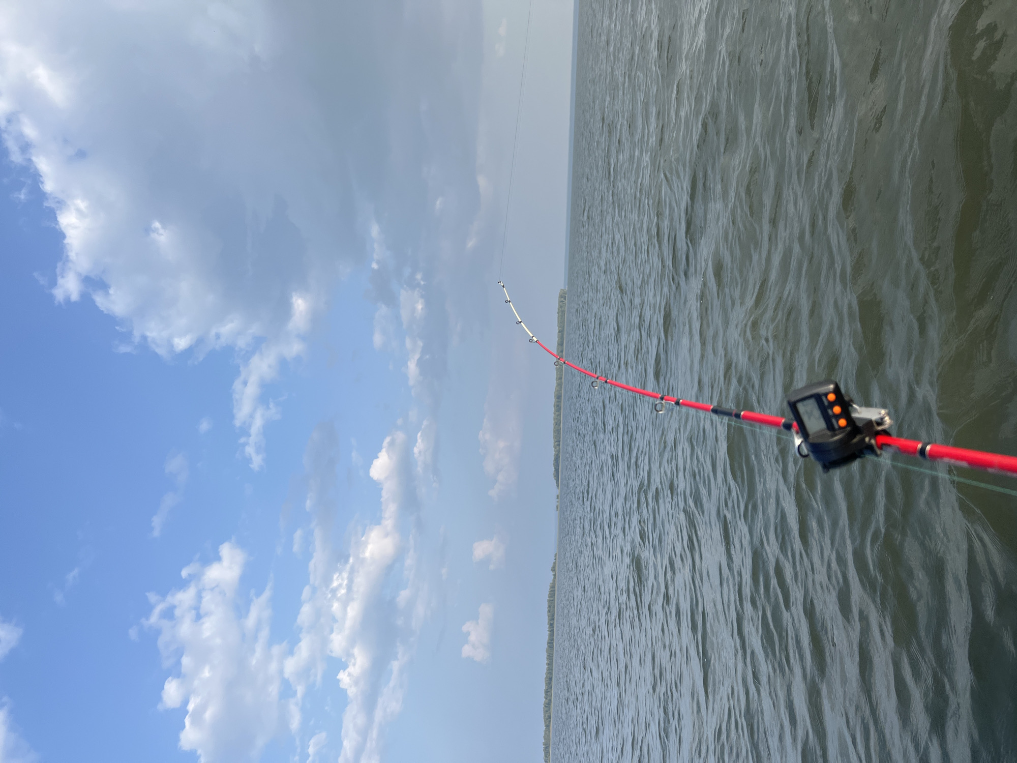 The image depicts a body of water, likely a lake or river, with a fishing rod in the foreground. The fishing rod is red and white, and a small digital device is attached to it. The water has moderate ripples, suggesting a gentle breeze. In the distance, there are islands with trees, and the sky is blue with scattered white clouds. The scene appears to be set in a natural environment. No specific landmarks or buildings are visible to indicate a location within Novopichugovo, Russia.
