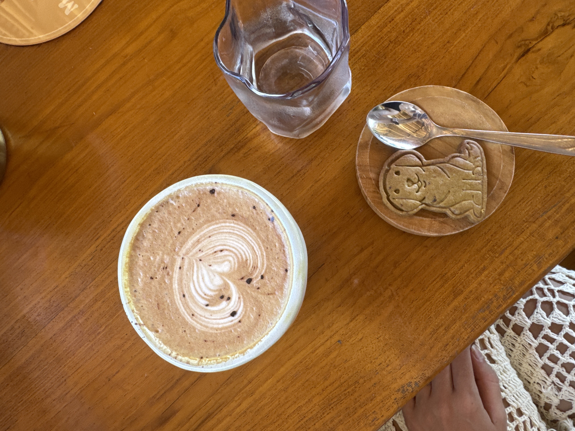 On a wooden table, a cup of cappuccino displays a latte art heart. Adjacent is a glass and a small wooden coaster with an animal-shaped cookie, over which a spoon is placed. A person wearing a white knitted garment is partially visible at the lower right. The location is Unknown, Unknown.