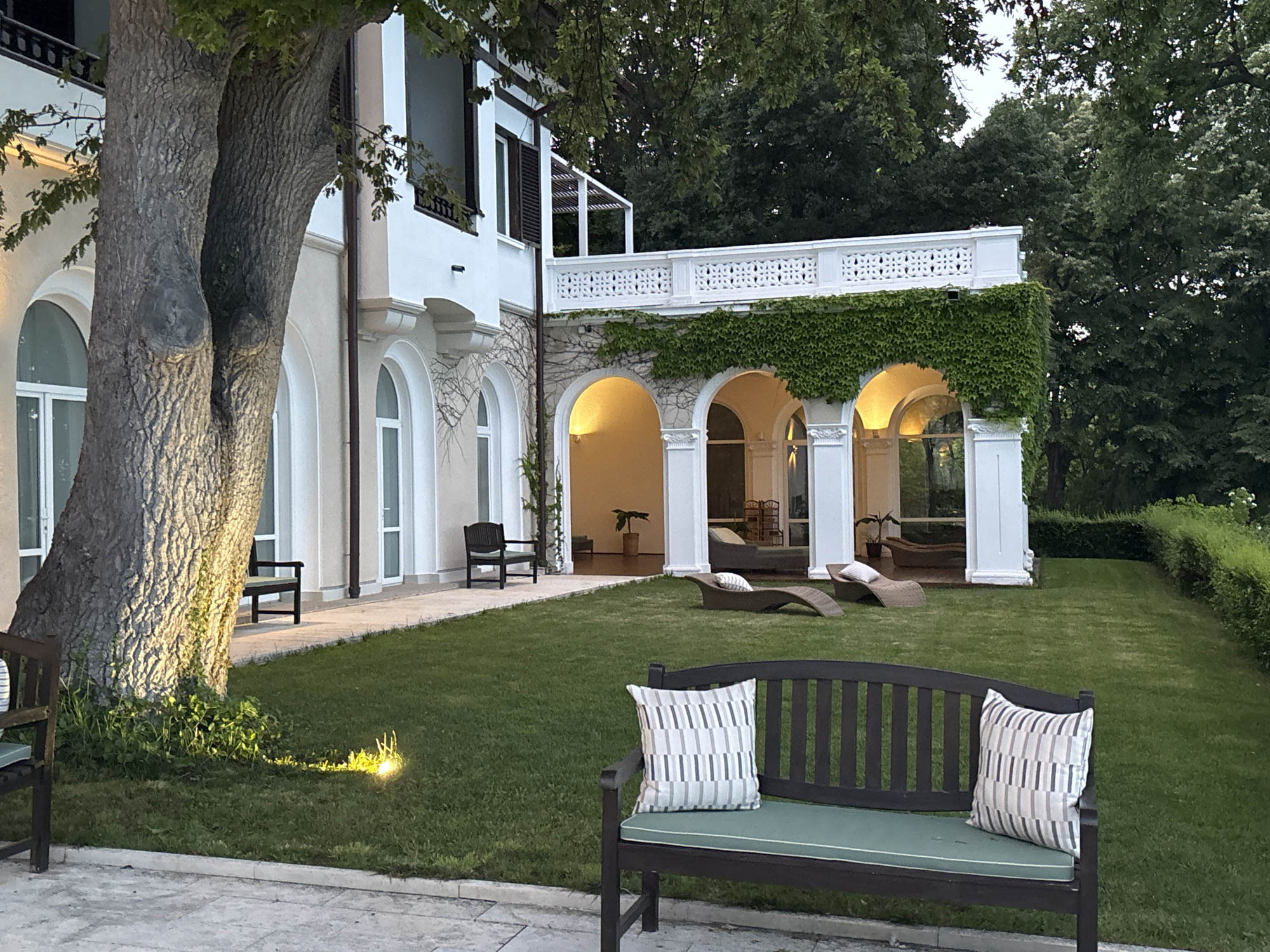 The image depicts the exterior of a villa, likely located in the city of Varna, Bulgaria, with a manicured lawn and surrounding trees. In the foreground, a dark wooden bench is positioned on a paved area, adorned with two decorative pillows featuring a striped pattern of grey and white. The bench faces a well-maintained green lawn. To the left of the bench, a large tree trunk with textured bark is visible, illuminated by a spotlight on the ground. In the middle ground, a series of arched walkways with illuminated interiors leads to the villa's main structure. Two lounge chairs are placed on the lawn in front of these arches. The villa itself is painted white, with arched windows and a white balustrade along an upper-level balcony, which is partially covered by climbing ivy. Dark wooden chairs are also visible on the paved area adjacent to the villa. The overall scene suggests a tranquil, upscale outdoor setting, likely in the evening or at dusk, given the artificial lighting.