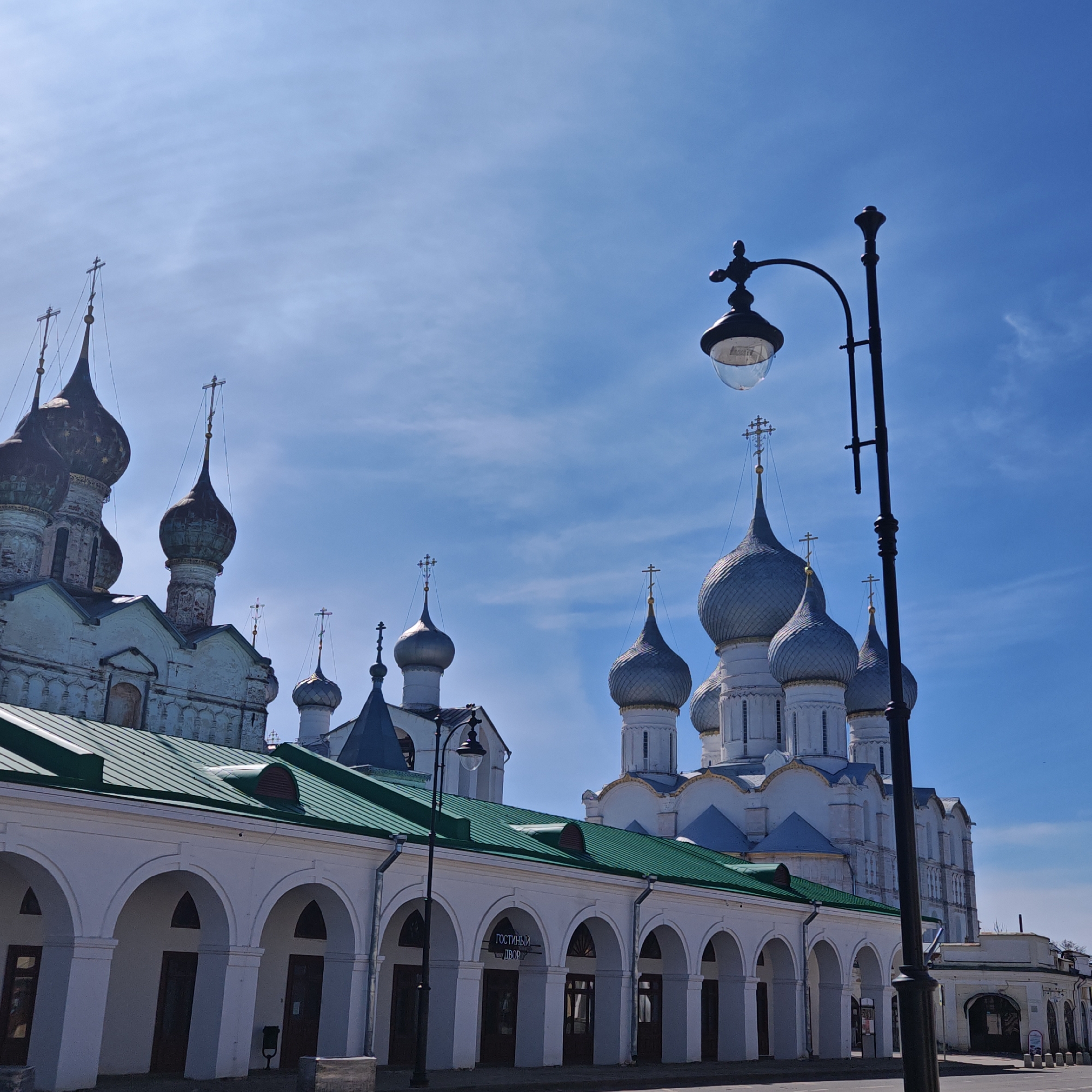 The image shows a sunny, daytime view of part of the Kremlin in Rostov, Russia. The architecture includes white buildings with green roofs, arched colonnades, and traditional Russian Orthodox church structures with multiple domes topped with crosses. One church has silver domes, another has dark domes. A tall, black street lamp is in the foreground on the right. Other, shorter street lamps are also visible along the colonnade. The sky is a bright blue with some wispy clouds. The scene appears to be a quiet street or plaza with no people visible in the immediate foreground. A sign in Cyrillic is visible on the building below the green roof.