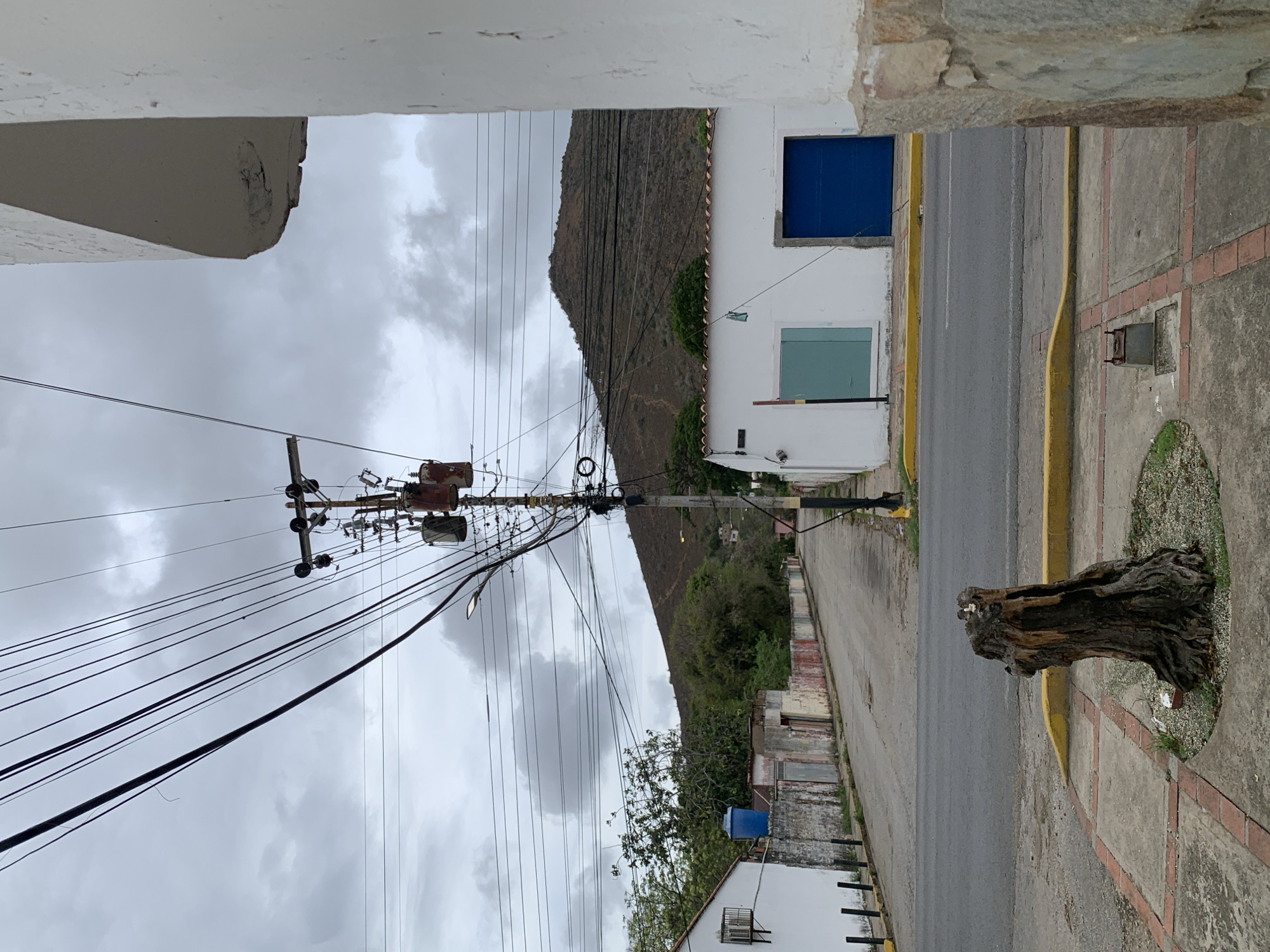 This image depicts a street scene in Santa Ana, Venezuela, under a heavily overcast, grey sky. A large, barren brown hill, sparsely dotted with green trees and bushes at its base, dominates the background.
An aged asphalt road curves gently uphill and to the left through the mid-ground. A tall utility pole stands centrally on the right side of the road, heavily laden with numerous black electrical wires running in multiple directions and supporting several rusted electrical transformers. A street light is also affixed to this pole, illuminating the road.
On the right side of the street, a single-story white building presents a bright blue garage door/shutter and a light green window/shutter, capped with a reddish-brown tiled roof. To its immediate right, a partial view of an older building shows an exposed stone and concrete wall. On the left side of the street, another single-story white building is visible, featuring an external air conditioning unit. Behind this, a low, multi-colored wall (faded pink, yellow, blue, and red) stretches along the street, with a blue water tank visible beyond it.
The foreground features a concrete sidewalk patterned with reddish-brown square tiles. A prominent, weathered, dark brown tree stump rests in a circular patch of gravel within the sidewalk in the center-left. A small, open metal utility access box is embedded in the sidewalk near the bottom right. Yellow painted curbs delineate the asphalt road from the sidewalk. The scene is devoid of people or vehicles.