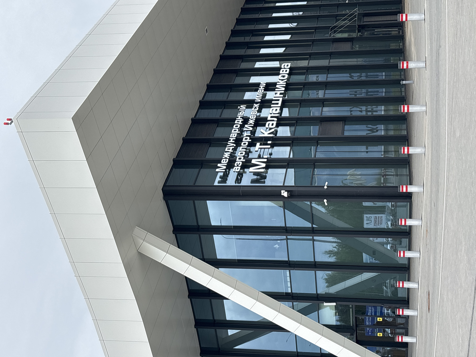 The image shows the exterior of a modern airport terminal building with large glass windows and a white, angular roof structure. White text on the building reads "Международный аэропорт Ижевск имени М.Т. Калашникова" which translates to "Izhevsk International Airport named after M.T. Kalashnikov." The airport is located in the village of Starye Martyanovo, Russia. In front of the building, there are several bollards with red and white stripes. The sky is overcast.