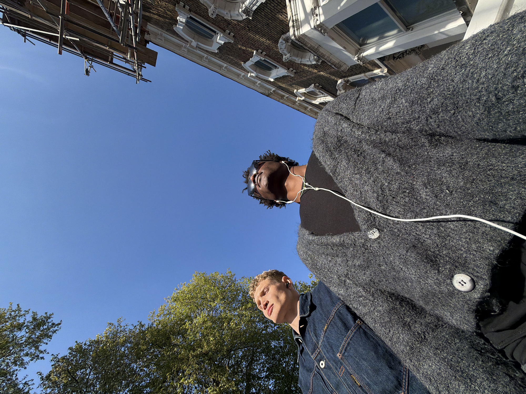 An upward-angled photograph, likely taken on a sunny day, captures two men against a bright blue sky with elements of an urban environment and nature. The perspective is from below, looking up.
In the foreground, slightly to the right, a man with light curly hair, appearing to be white, looks down and to his left. He is wearing a blue denim jacket with visible stitching and a small, unidentifiable gold-colored tag on his left breast pocket.
Behind him and slightly to the left, a second man, appearing to be Black, with dark dreadlocks or curly hair, looks upwards. He wears dark sunglasses and a plain black t-shirt underneath a thick, textured, dark grey cardigan. White wired earbuds are visible around his neck and in his ears. The cardigan has large, light grey buttons, one of which appears to have an indistinct "11" or "II" engraved or embossed on it.
The background is dominated by a clear, bright blue sky, indicating good weather and strong sunshine. In the upper right corner, an ornate building with multiple windows and decorative white architectural elements is partially visible. Scaffolding is attached to the upper section of this building, suggesting construction or renovation, a common sight in London. In the lower left, the green foliage of trees is visible, suggesting a park or tree-lined street.
The scene depicts two individuals, possibly friends, walking or pausing outdoors in what appears to be an urban setting, likely London given the architectural style and general environment. There is no clear activity other than their presence and differing gazes.